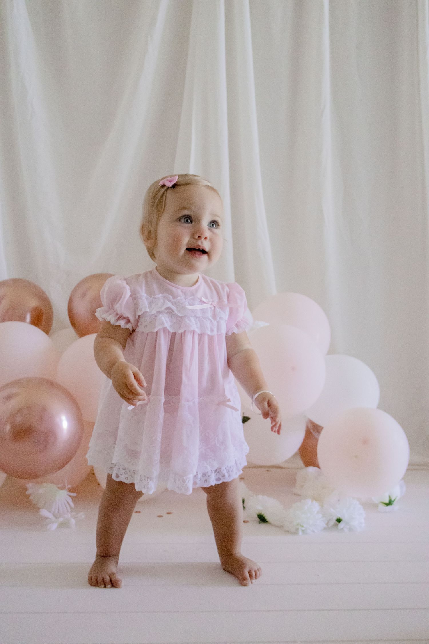 Baby standing in pink lace dress with rose gold and white balloons for first birthday photoshoot.