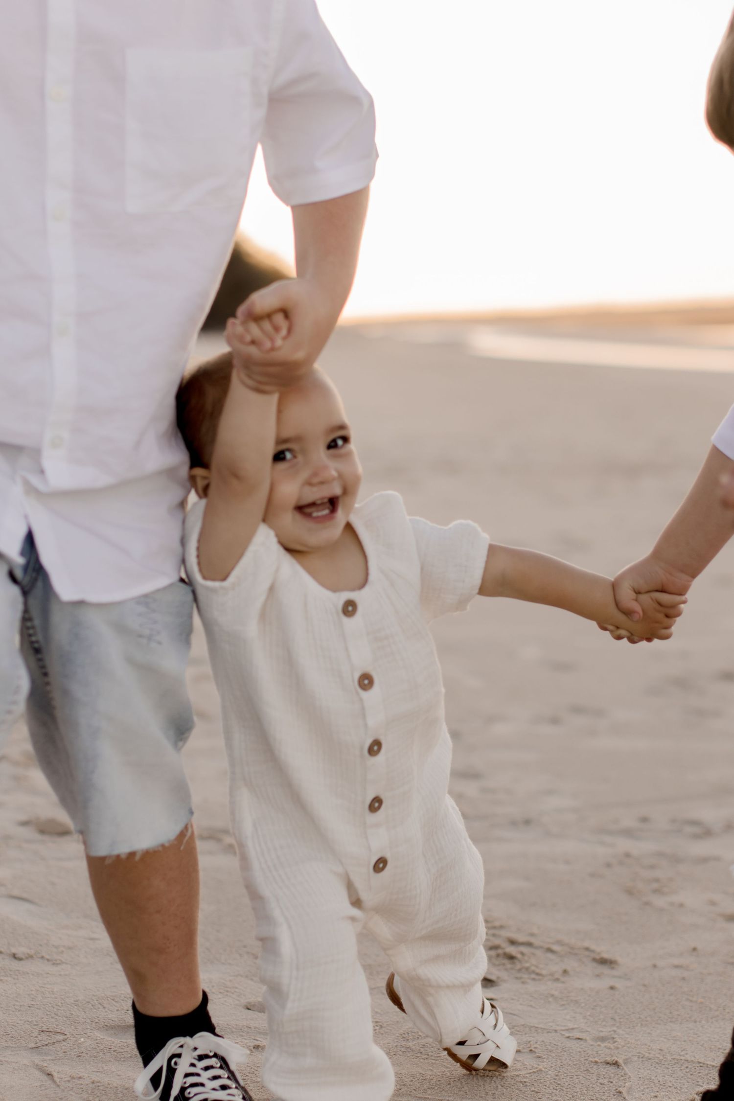 A small child in a white button-up romper holding hands with parents on a sandy beach at sunset.