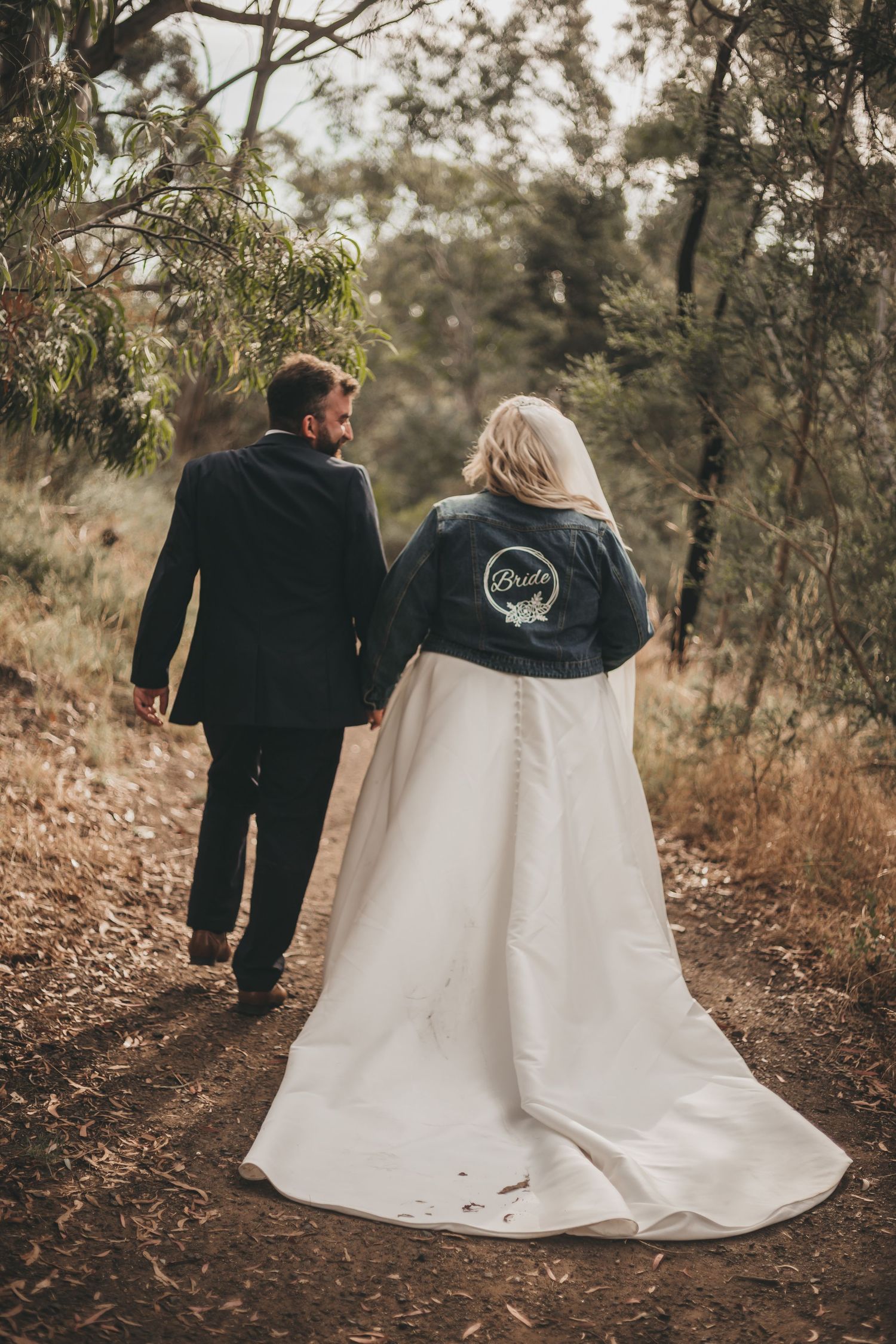A couple walks down a forest path on their wedding day, with the bride wearing a white gown and denim jacket.