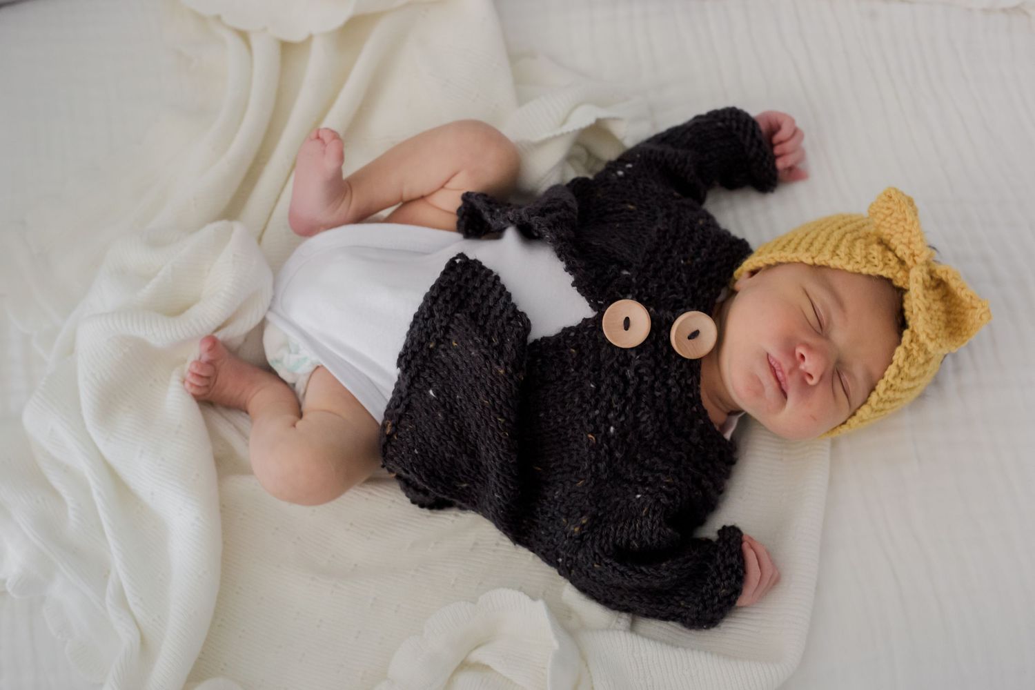Baby in black knit cardigan and yellow beanie resting on white bed linens.
