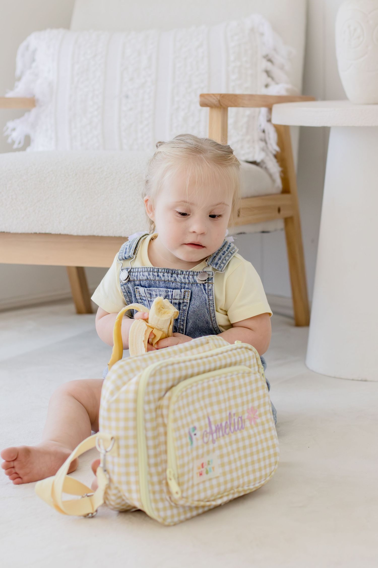 Toddler girl with Down syndrome sits on floor exploring a yellow gingham personalized backpack named Amelia.