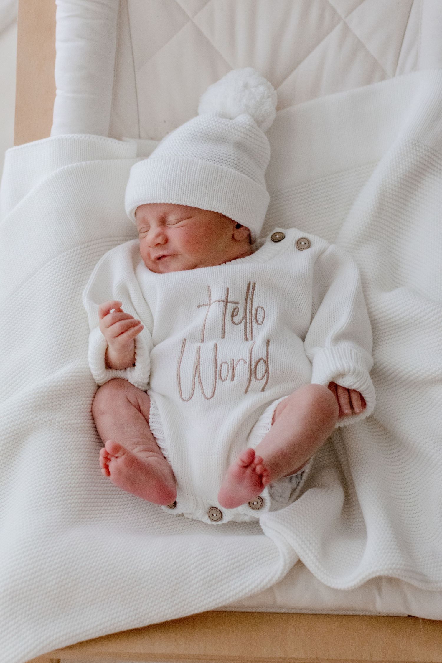 Newborn baby wearing white knit hat and 'Hello World' onesie sleeping peacefully on white blanket.