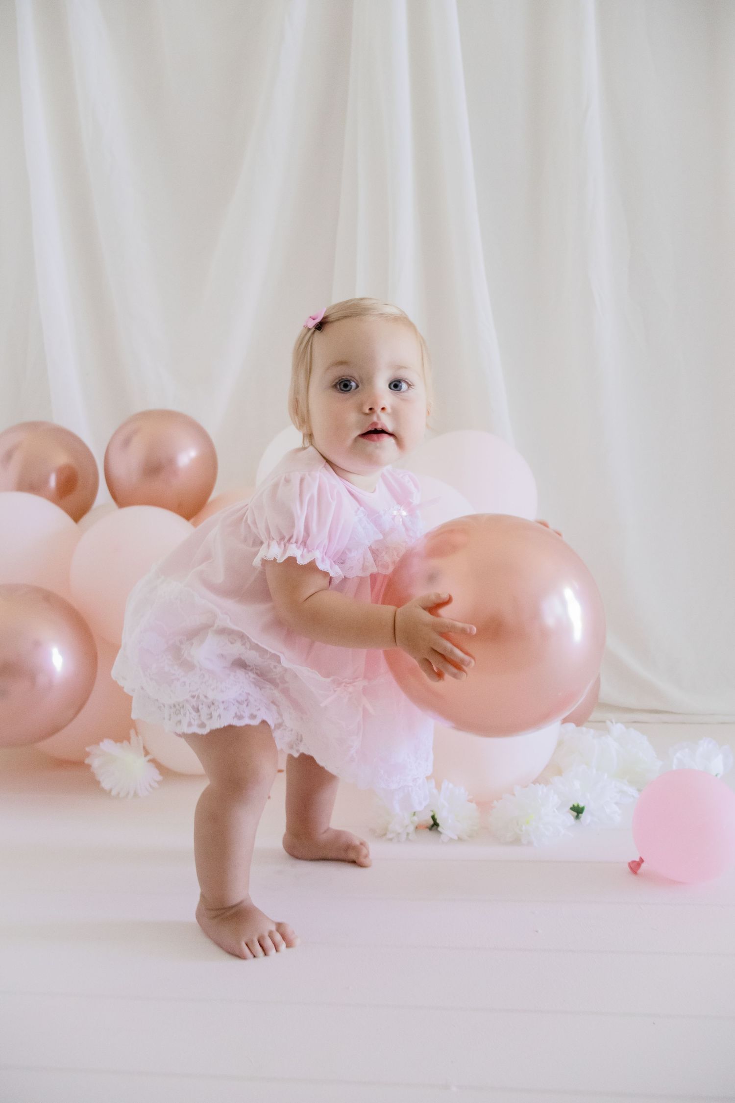Toddler in delicate pink dress playing with rose gold and white balloons against white backdrop.