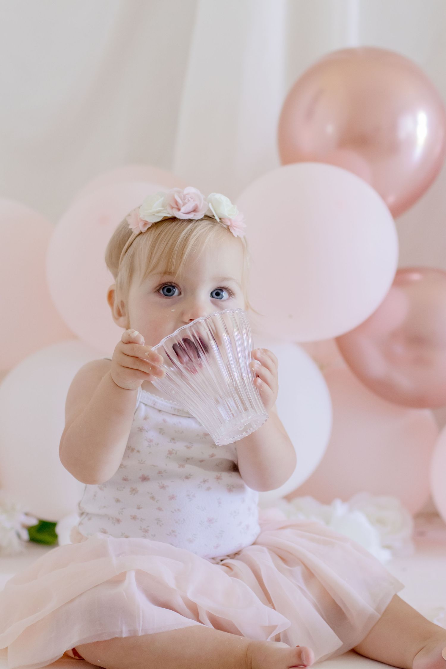 Baby wearing floral crown peers through crystal glass with pink balloons behind.