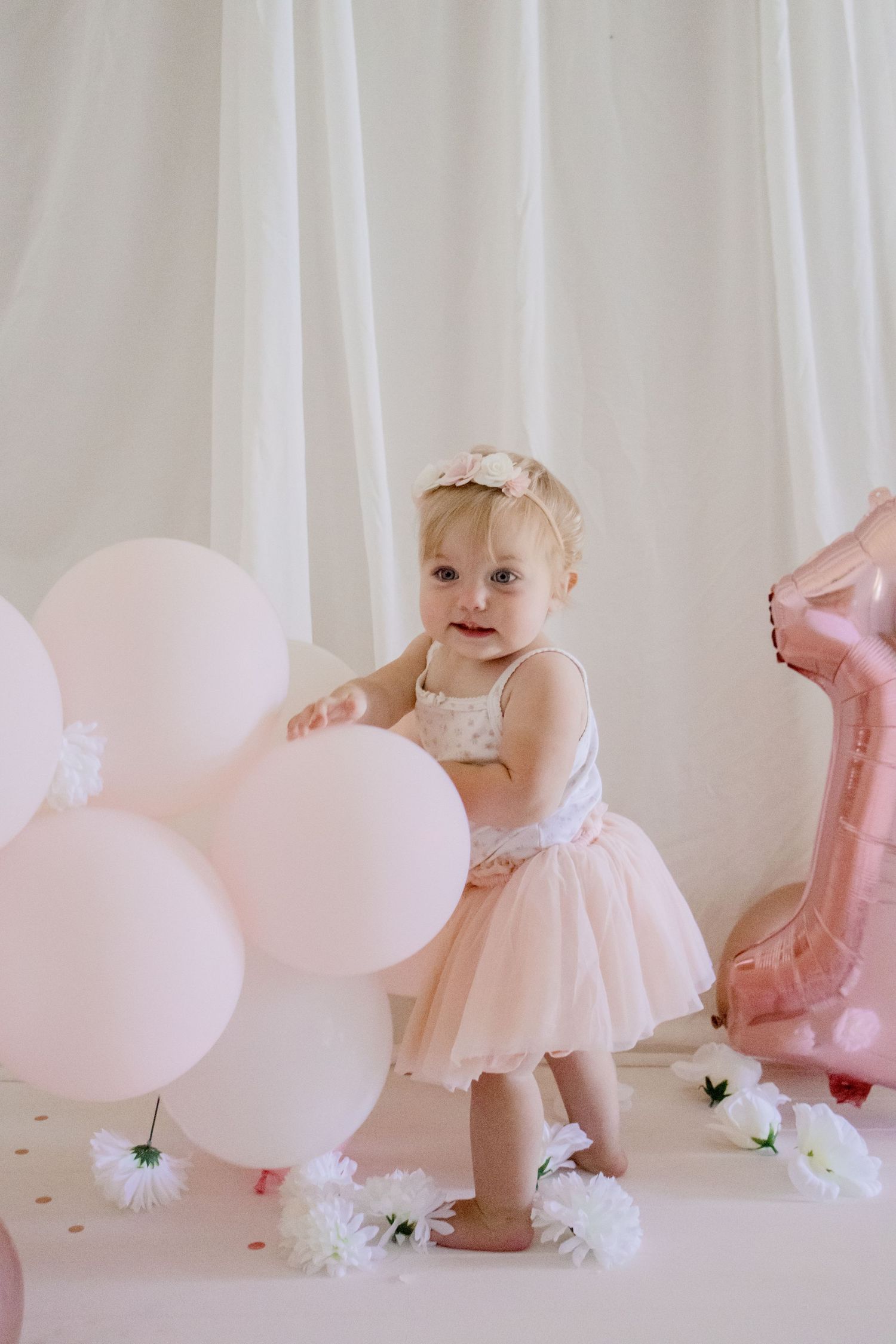 A small child in a festive dress plays among pink balloons and white flowers at a photo shoot.