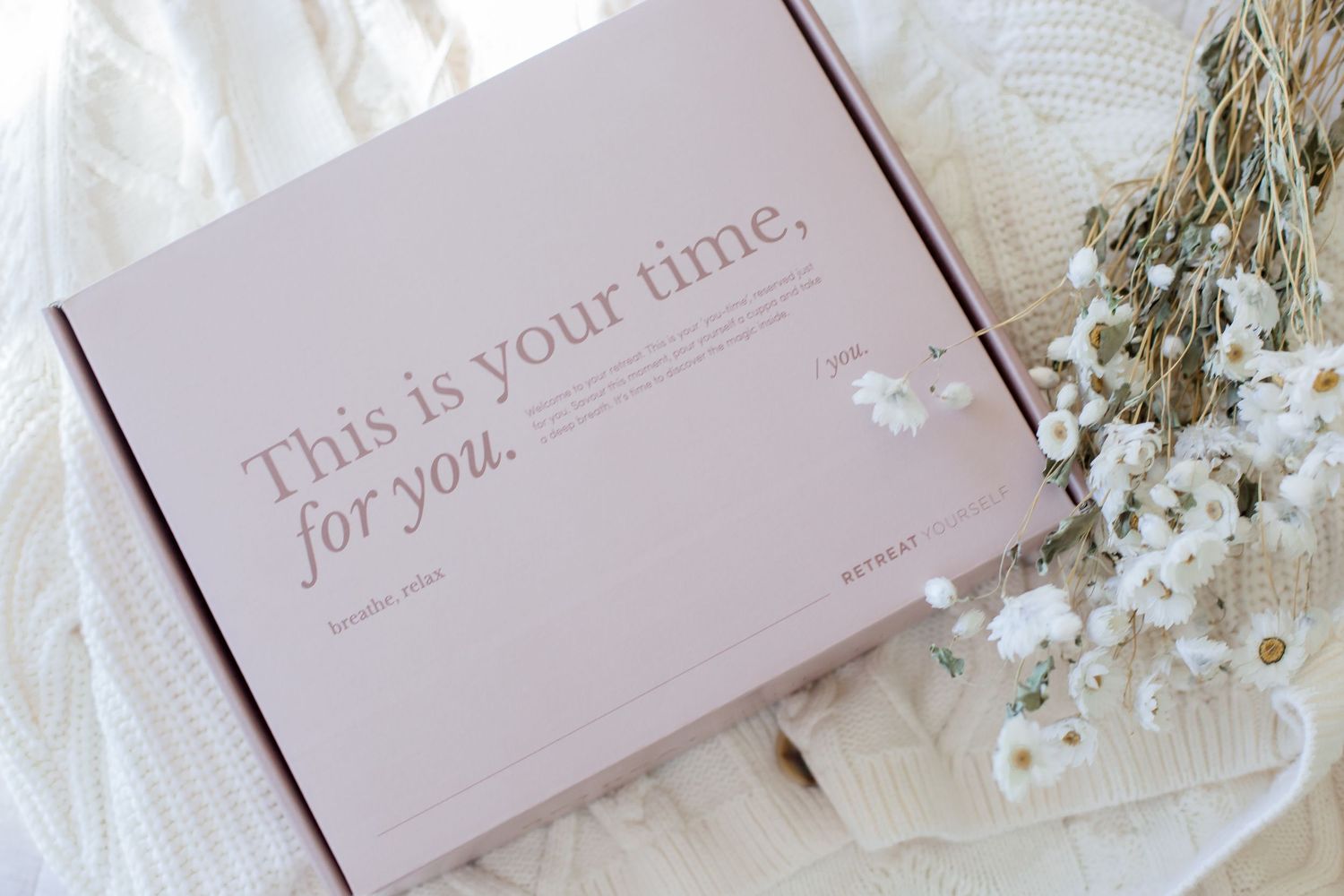 A pink gift box with text 'This is your time for you' on a white fabric background with dried flowers.