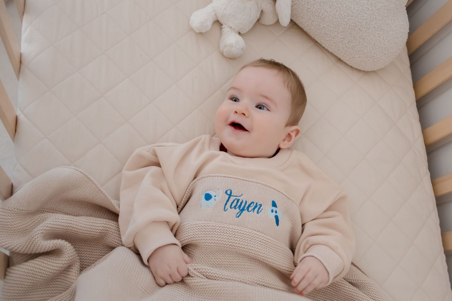 Baby wearing beige sweater with blue text lying on cream-colored bedding in a crib.