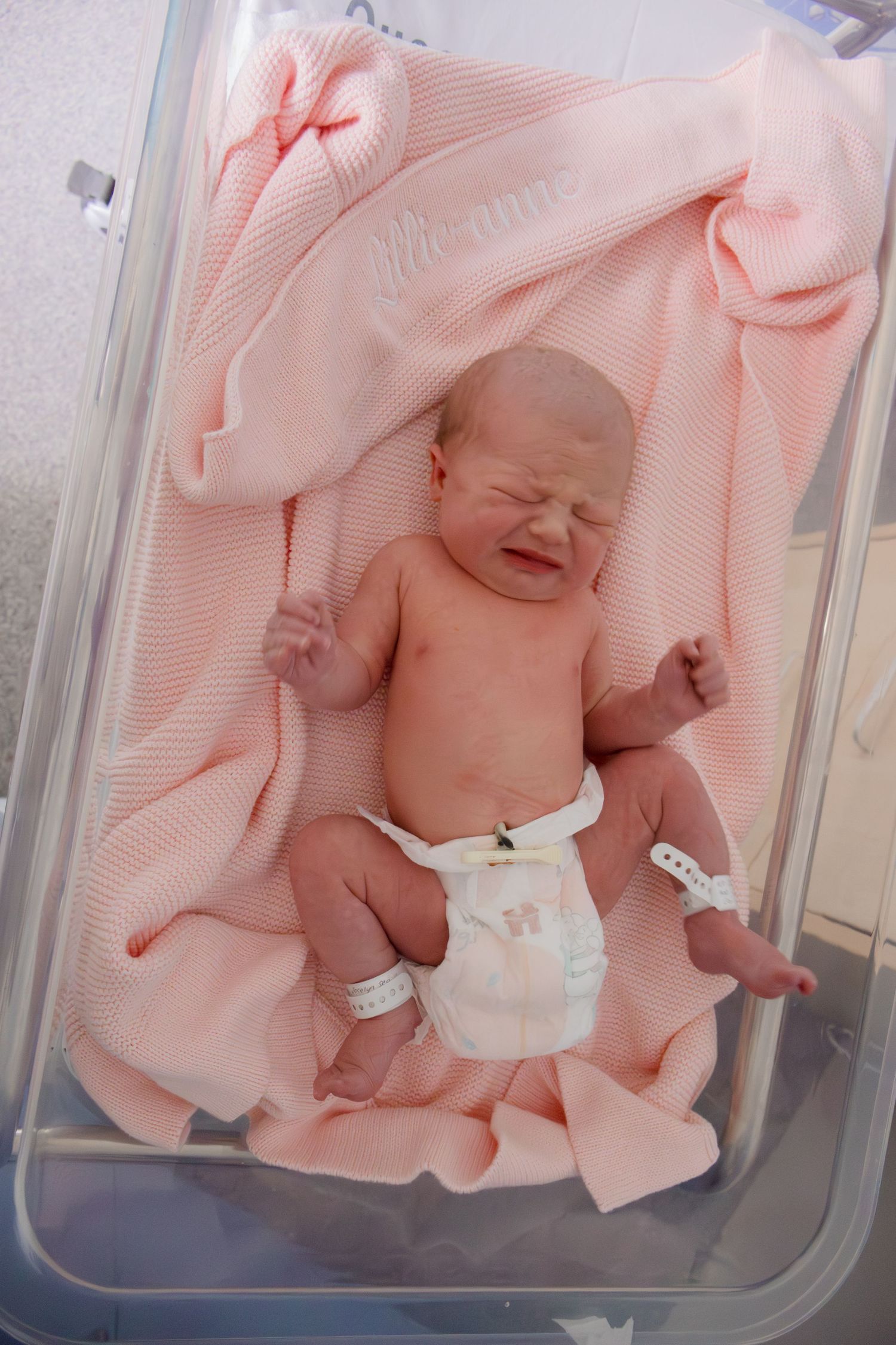 Crying newborn baby in hospital bassinet wrapped in pink blanket with ID bracelet.