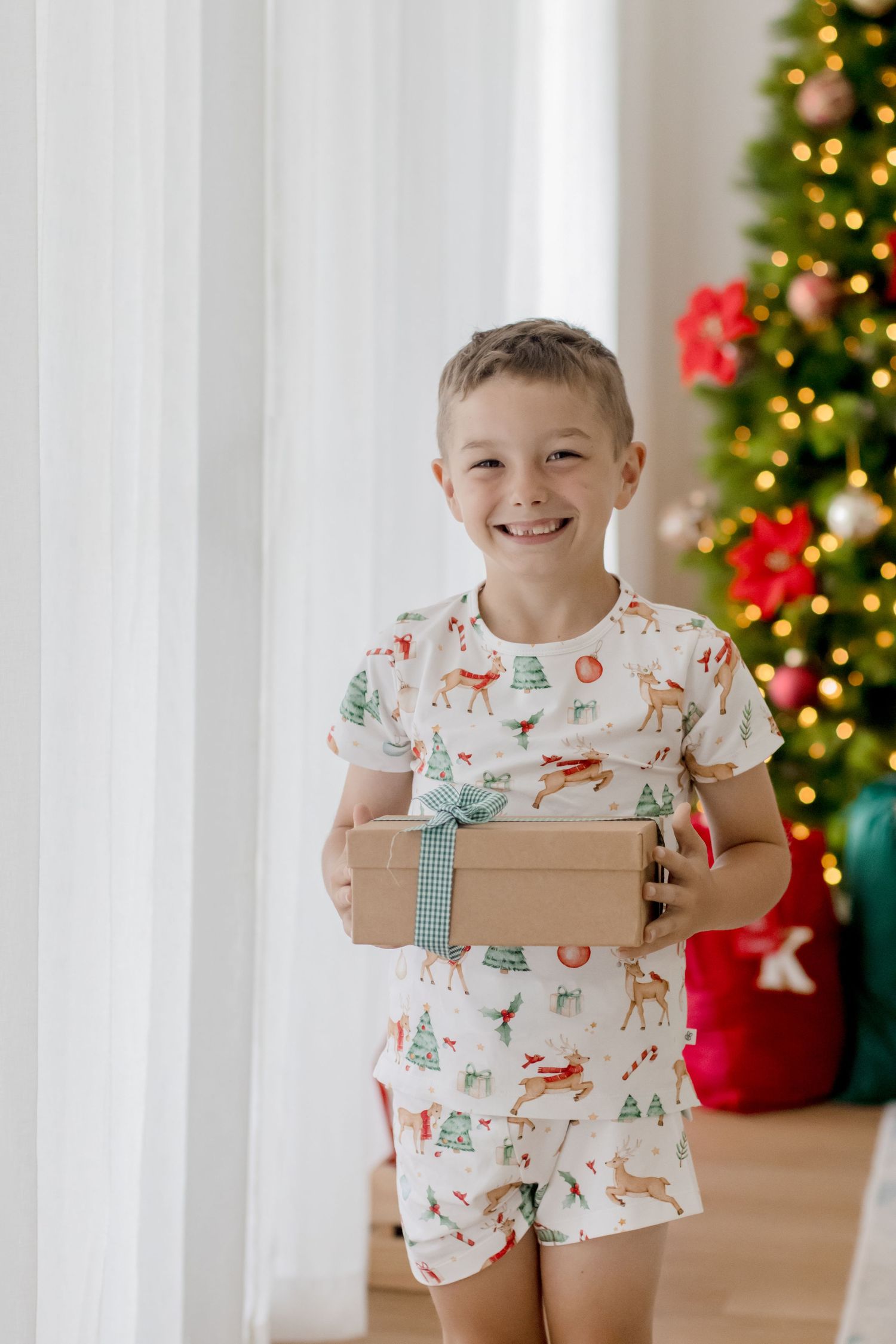 Child wearing holiday pajamas holds a brown box while standing in front of decorated Christmas trees.