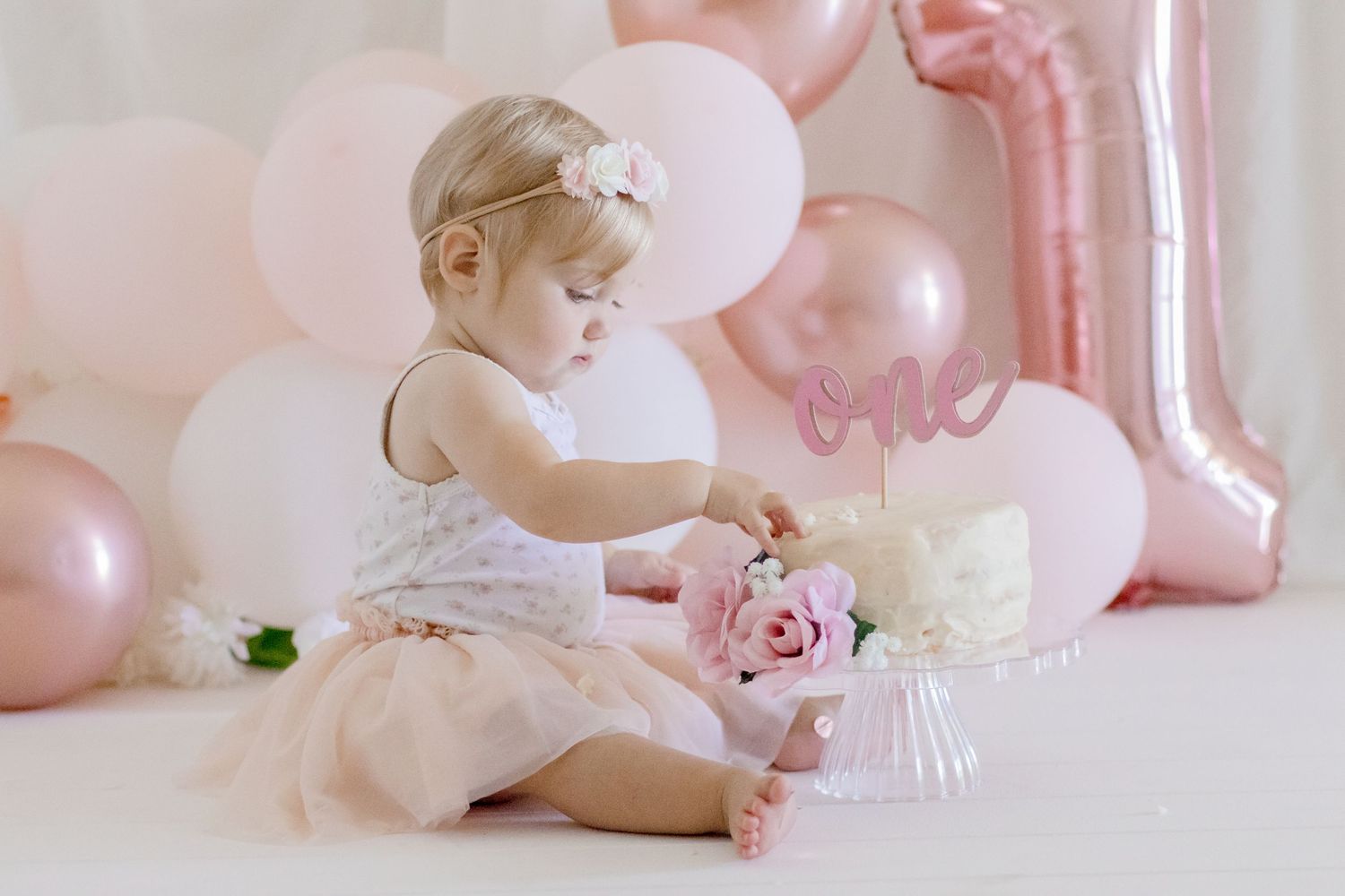 A baby in white dress reaching for a decorated cake against pink balloon backdrop.