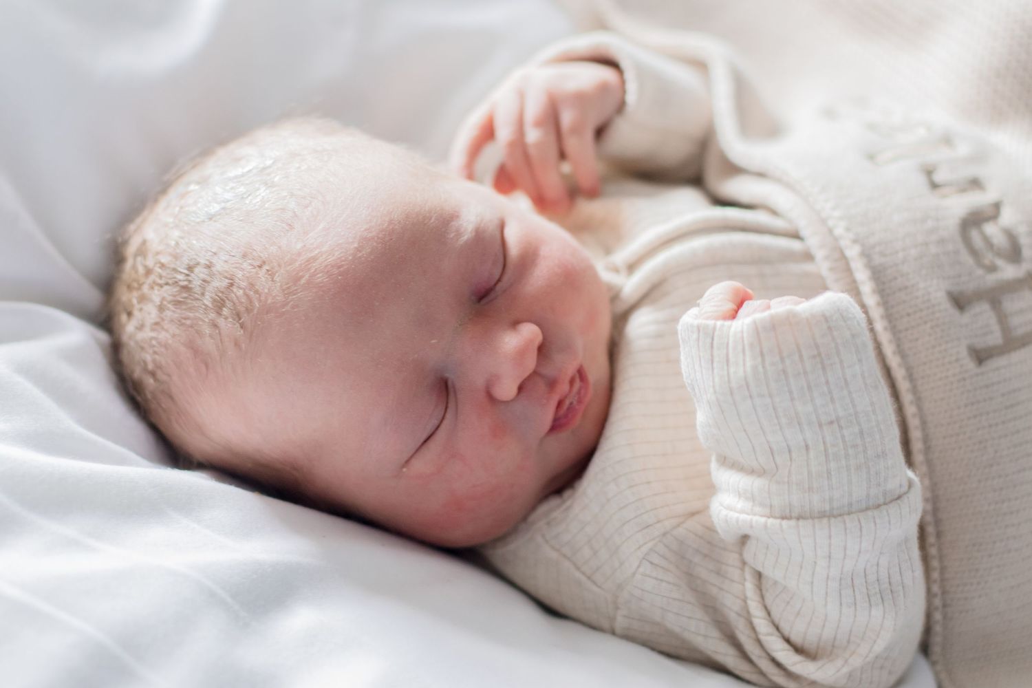 A sleeping newborn in a cream colored sweater resting peacefully on white bedding.