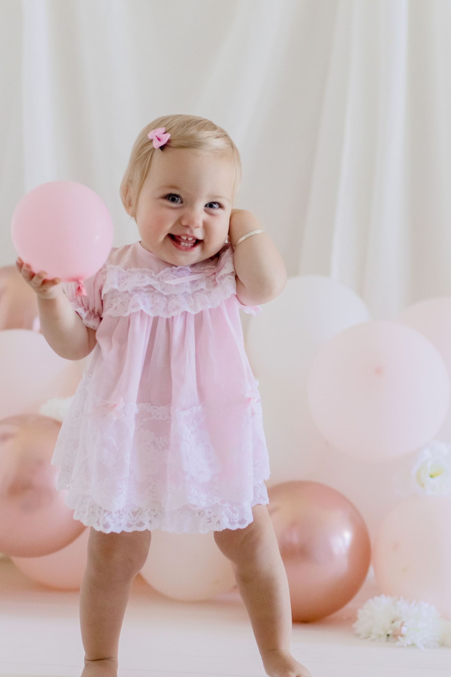 Playful toddler in lace dress holds pink balloon during photo session.
