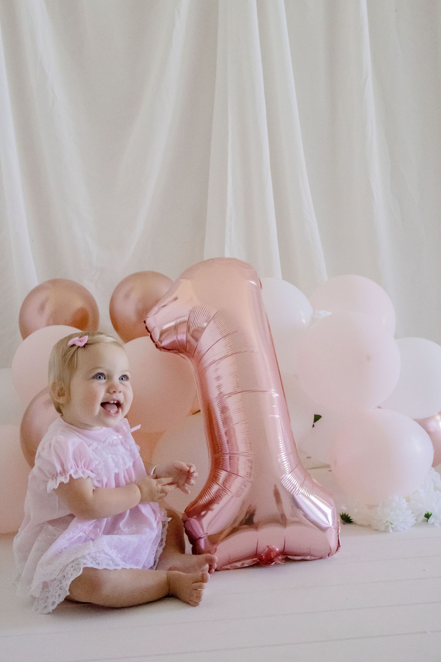 Baby in pink dress sitting next to rose gold number one balloon and pastel balloons against white backdrop.