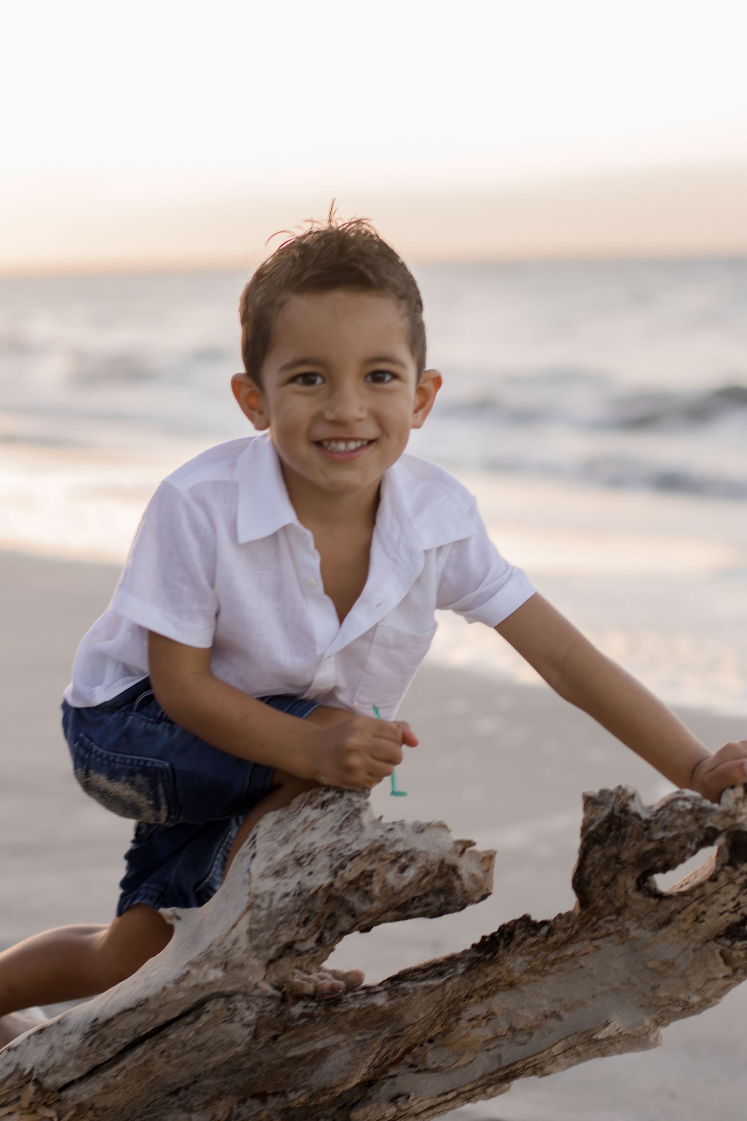 A child sits on driftwood at the beach during sunset, wearing a white shirt and denim shorts.