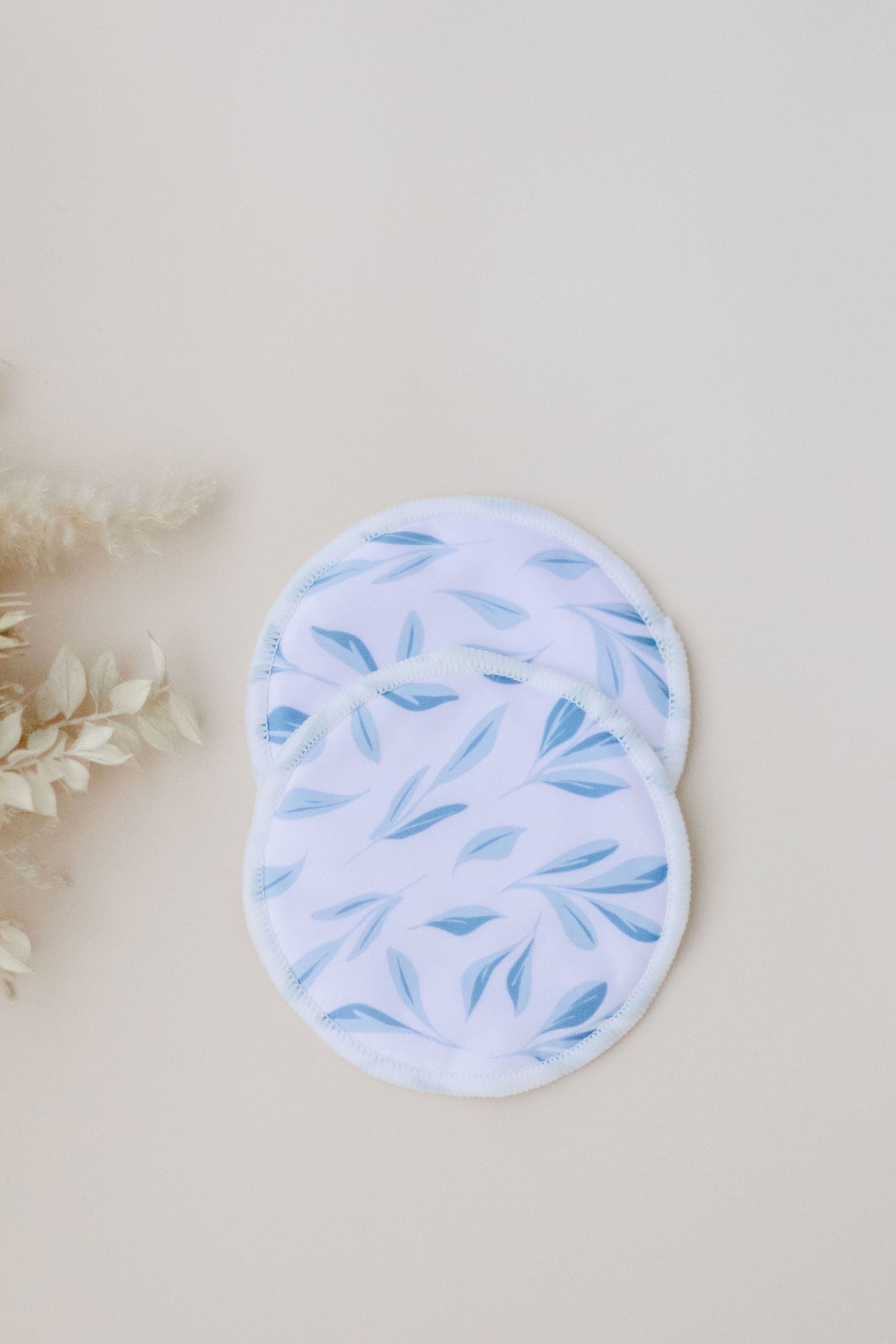 Two white fabric nursing pads with delicate blue floral leaf pattern designs on a neutral beige background.