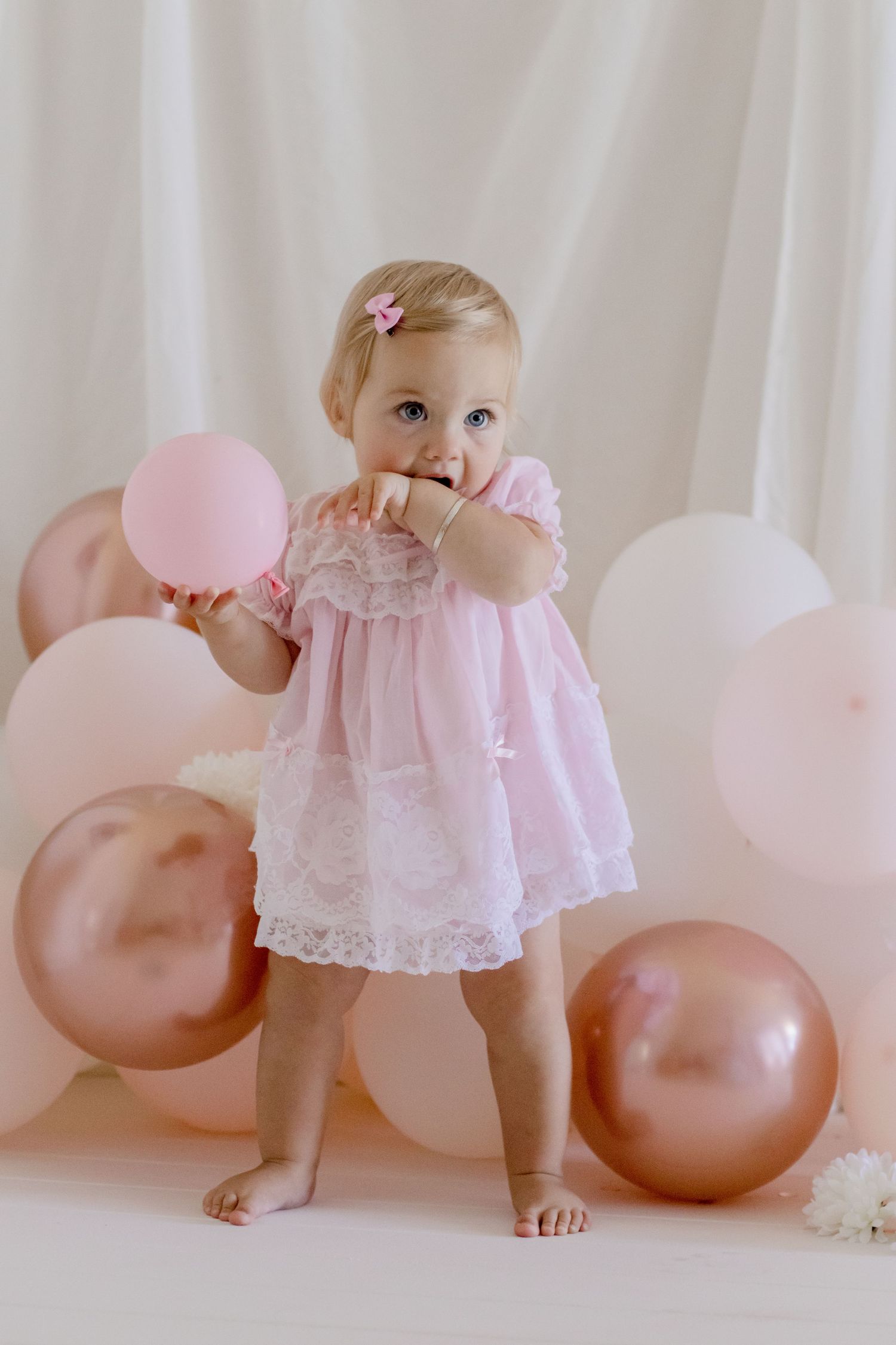 Baby in pink lace dress plays with balloons during first birthday photoshoot.