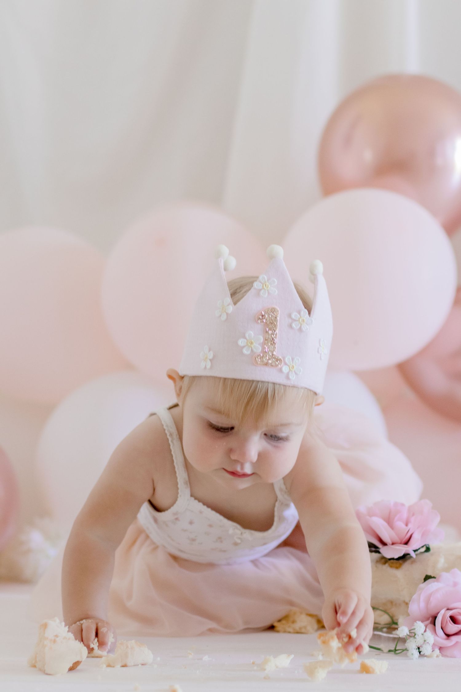 A toddler in crown and dress reaching for party decorations on white table.