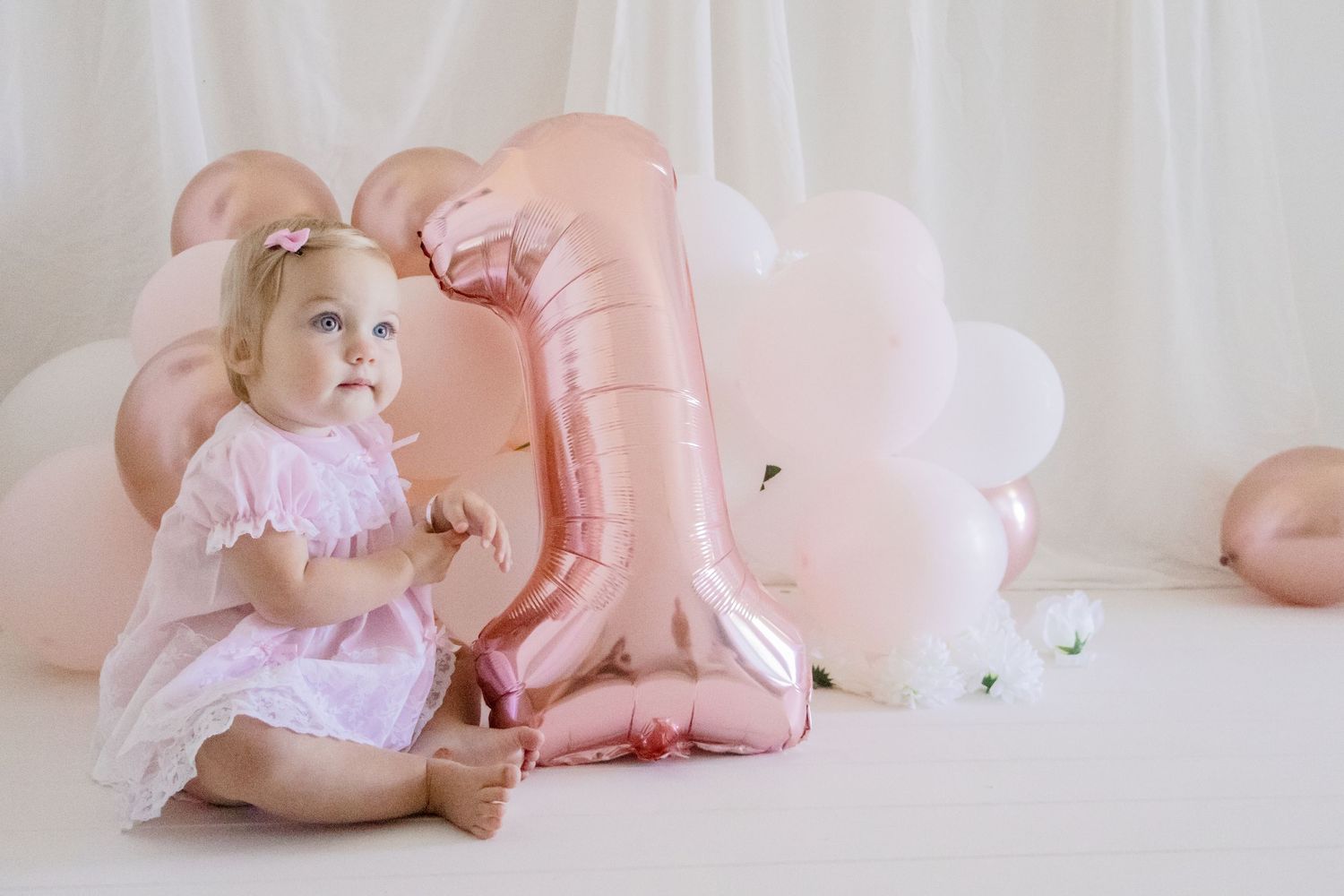 Excited baby in pink dress beside large metallic number one balloon with white backdrop.