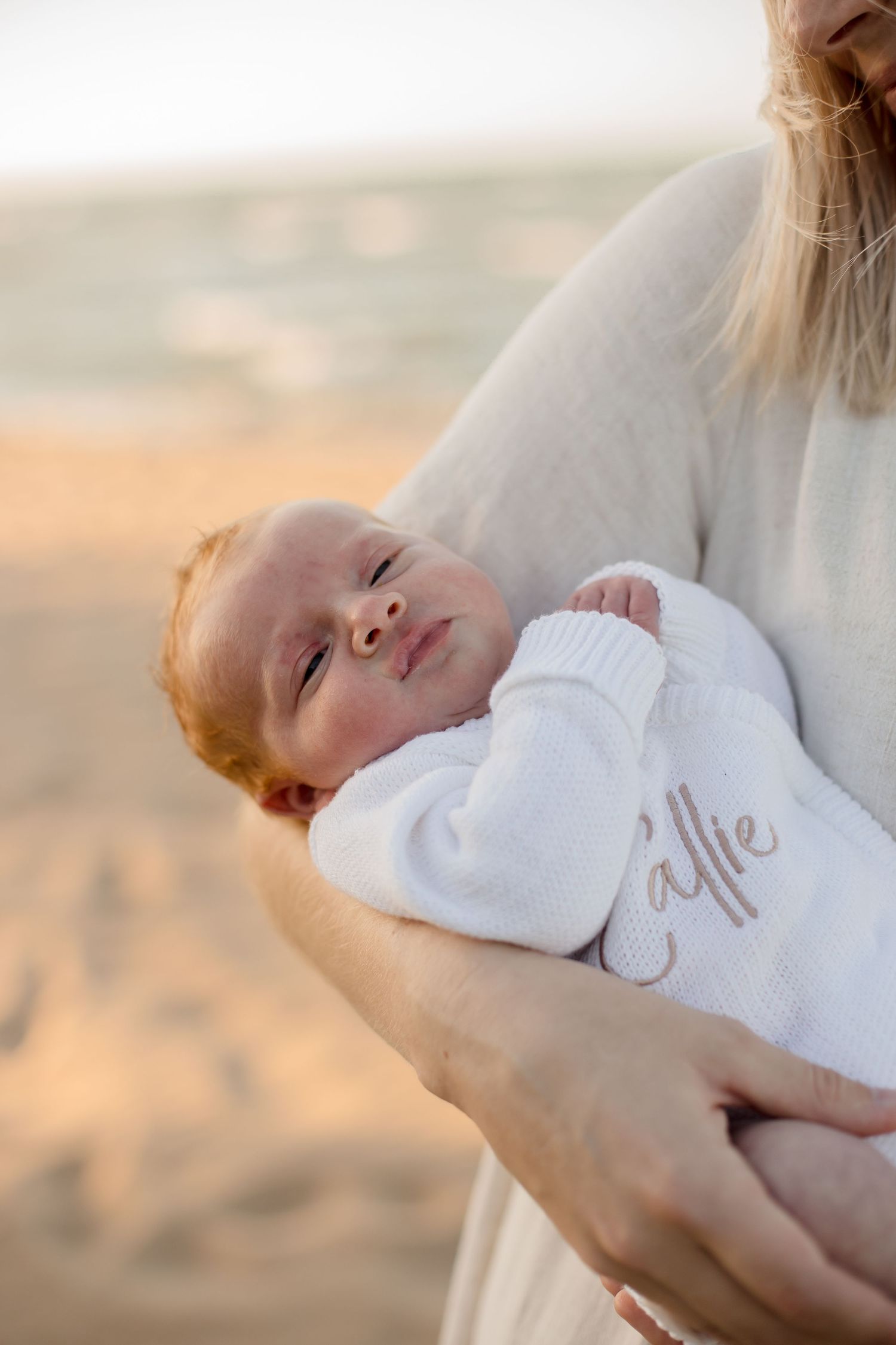 Newborn in white outfit smiles while being held against sunset beach backdrop.
