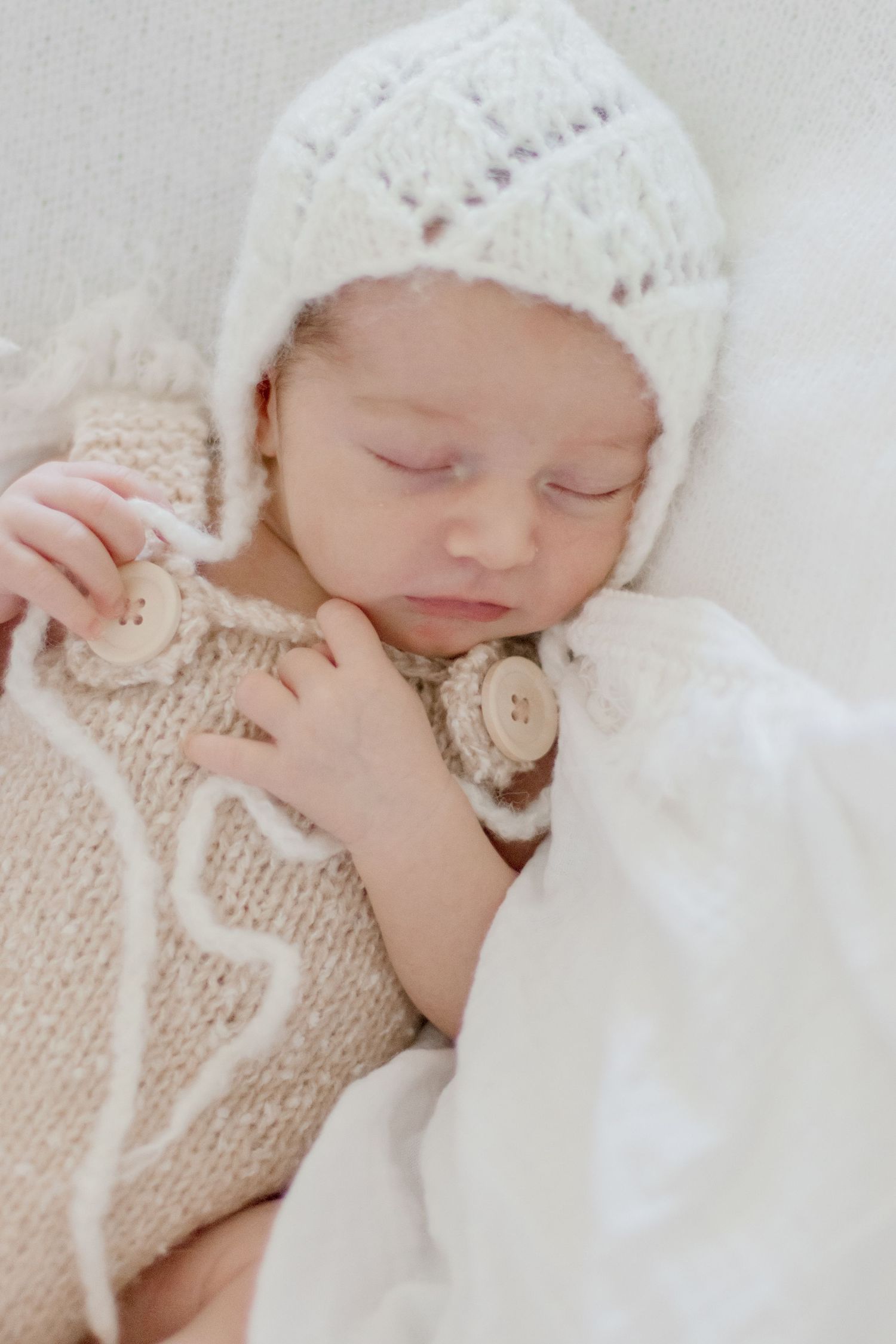 Sleeping newborn in knit white bonnet and sweater cuddles soft blanket.