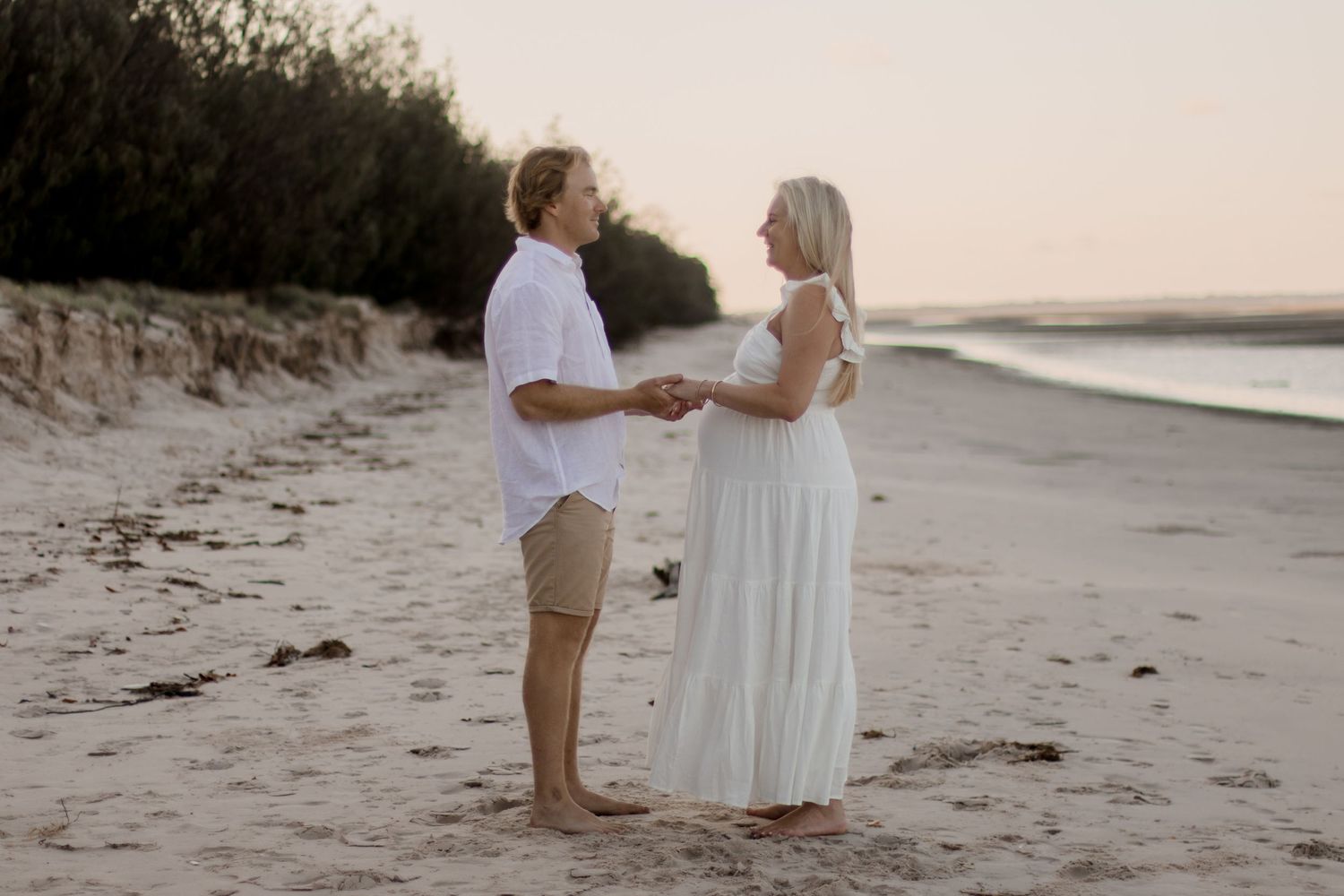 A couple holds hands during their beachside maternity photoshoot at dusk.