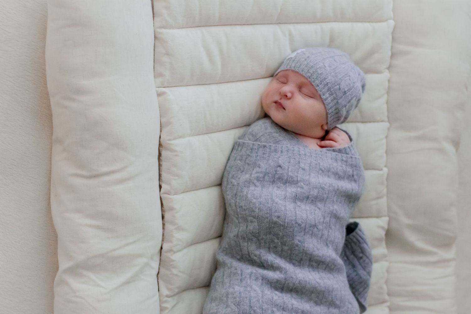 A sleeping baby wearing a gray knitted outfit and hat rests on a plush white quilted surface.