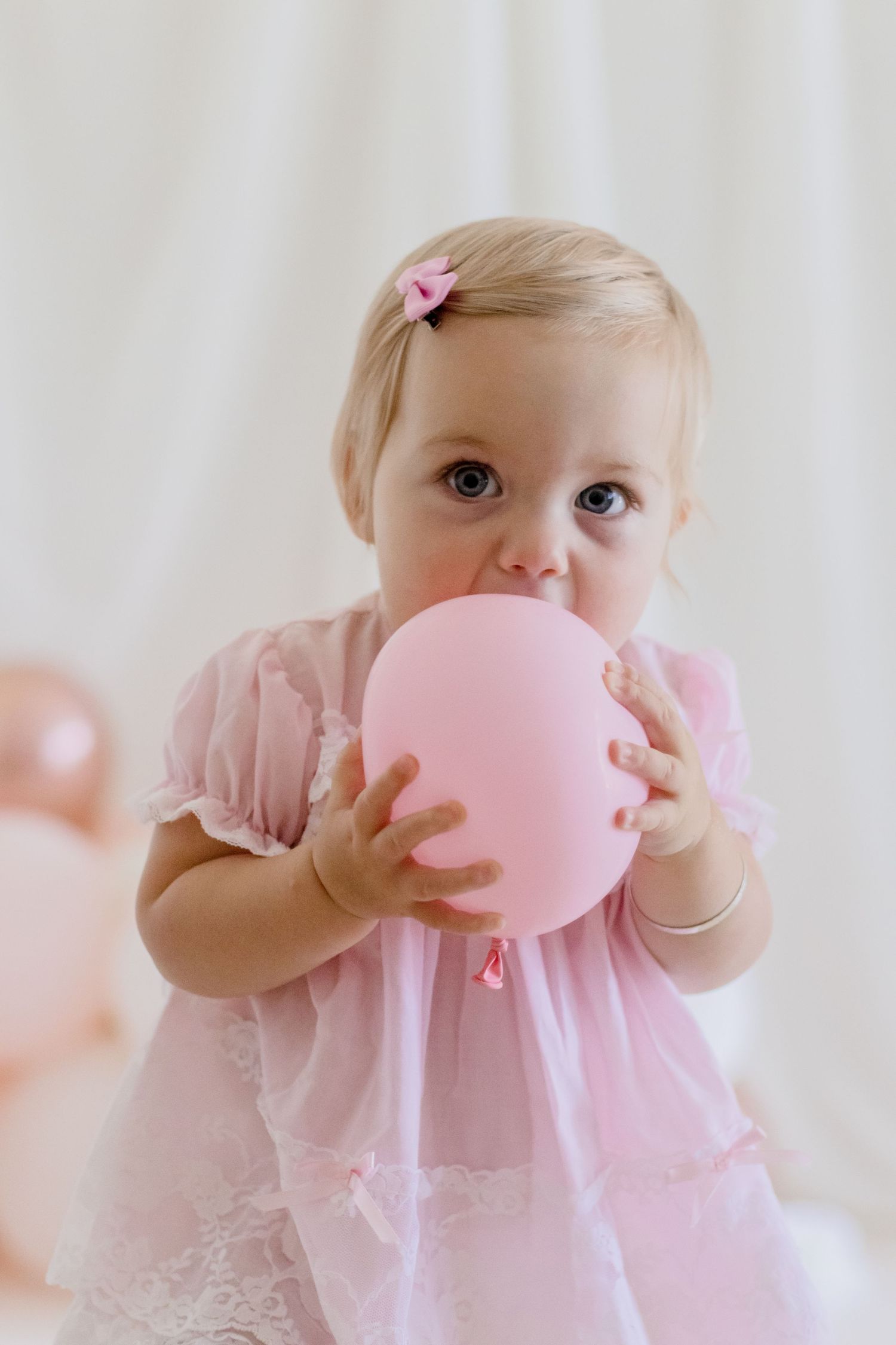 Toddler in pink dress blowing up pink balloon against white background.