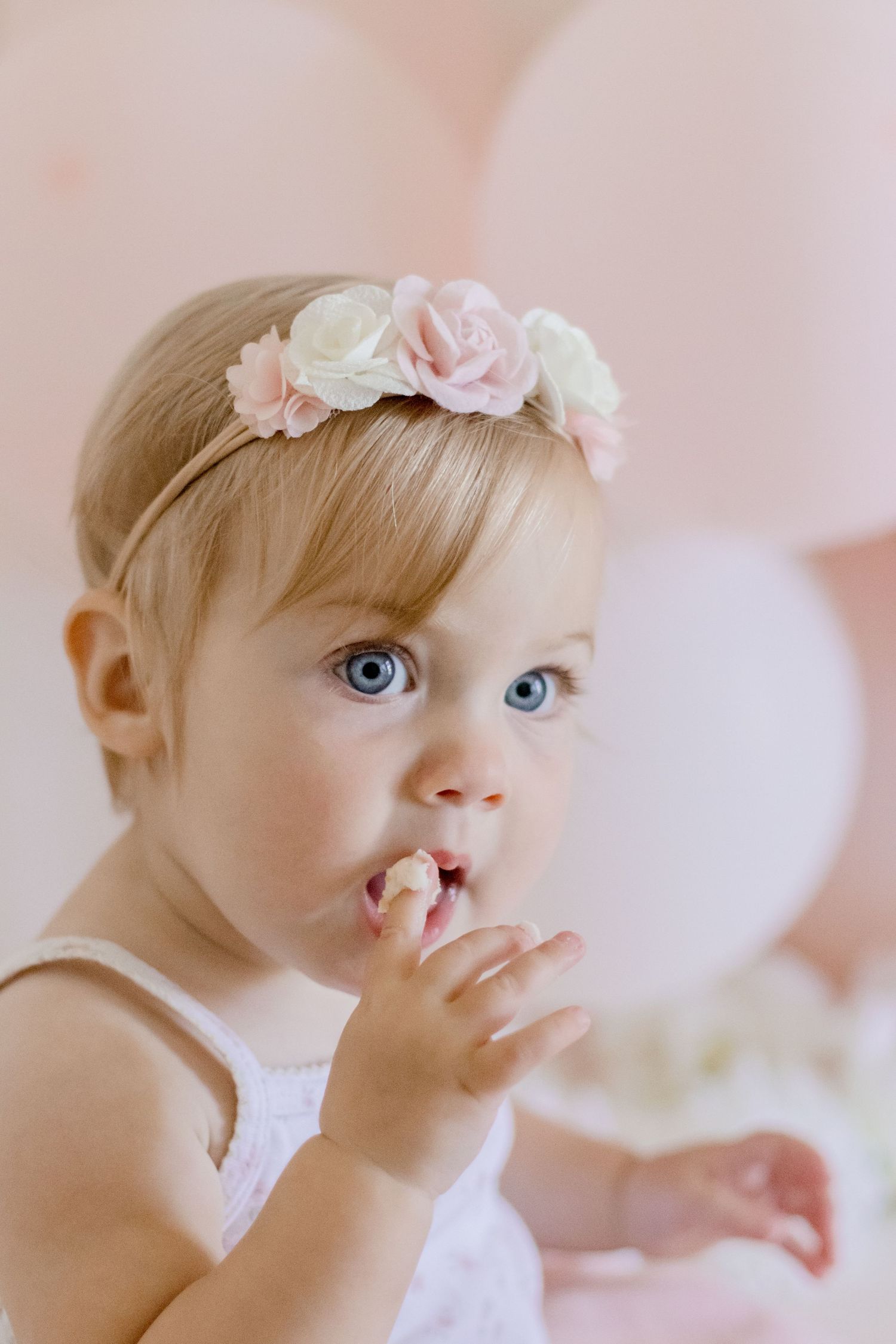 Baby wearing white and pink floral headband with bright blue eyes and finger in mouth against pink background.