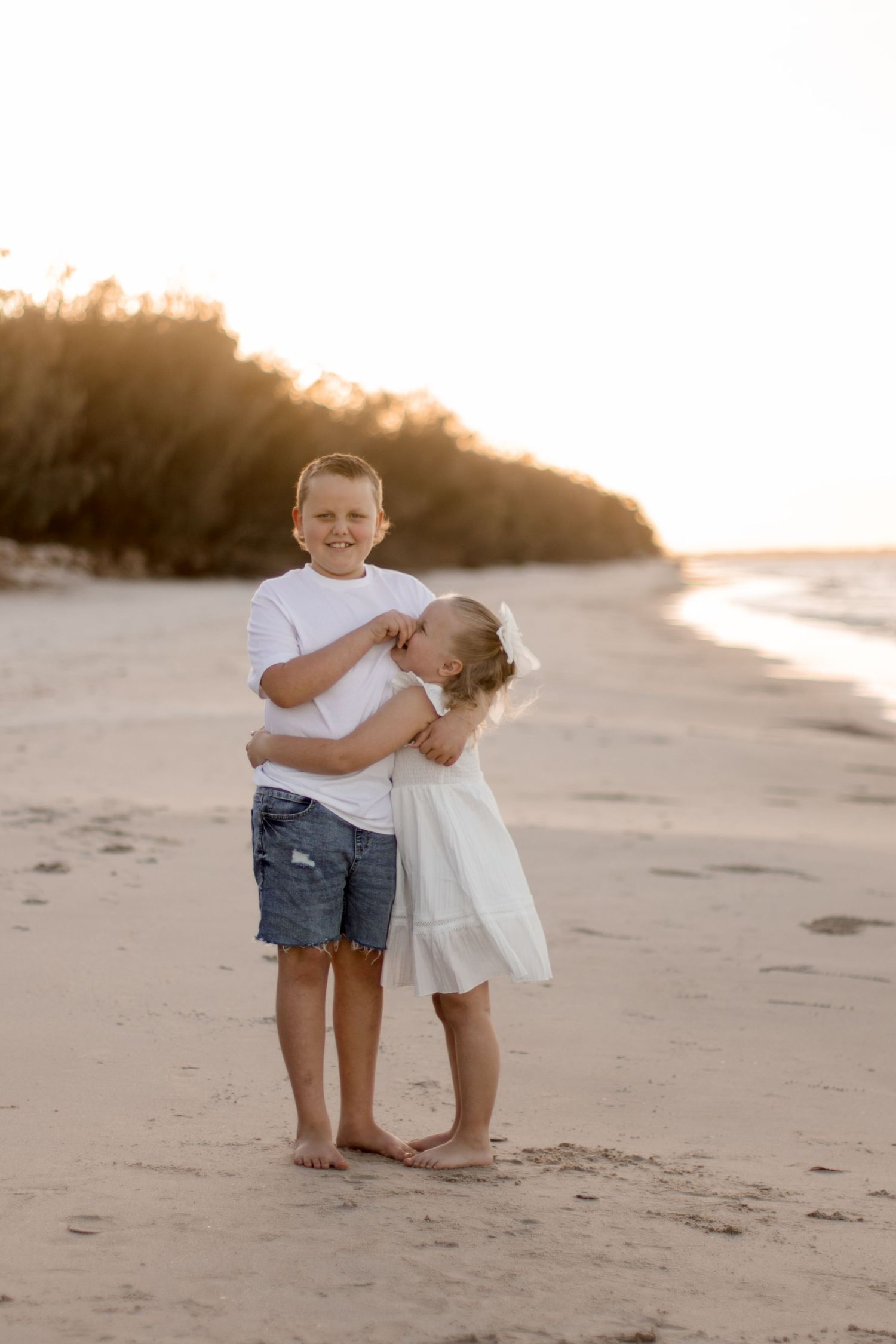 Two children stand close together on a beach at sunset with trees visible in the background.