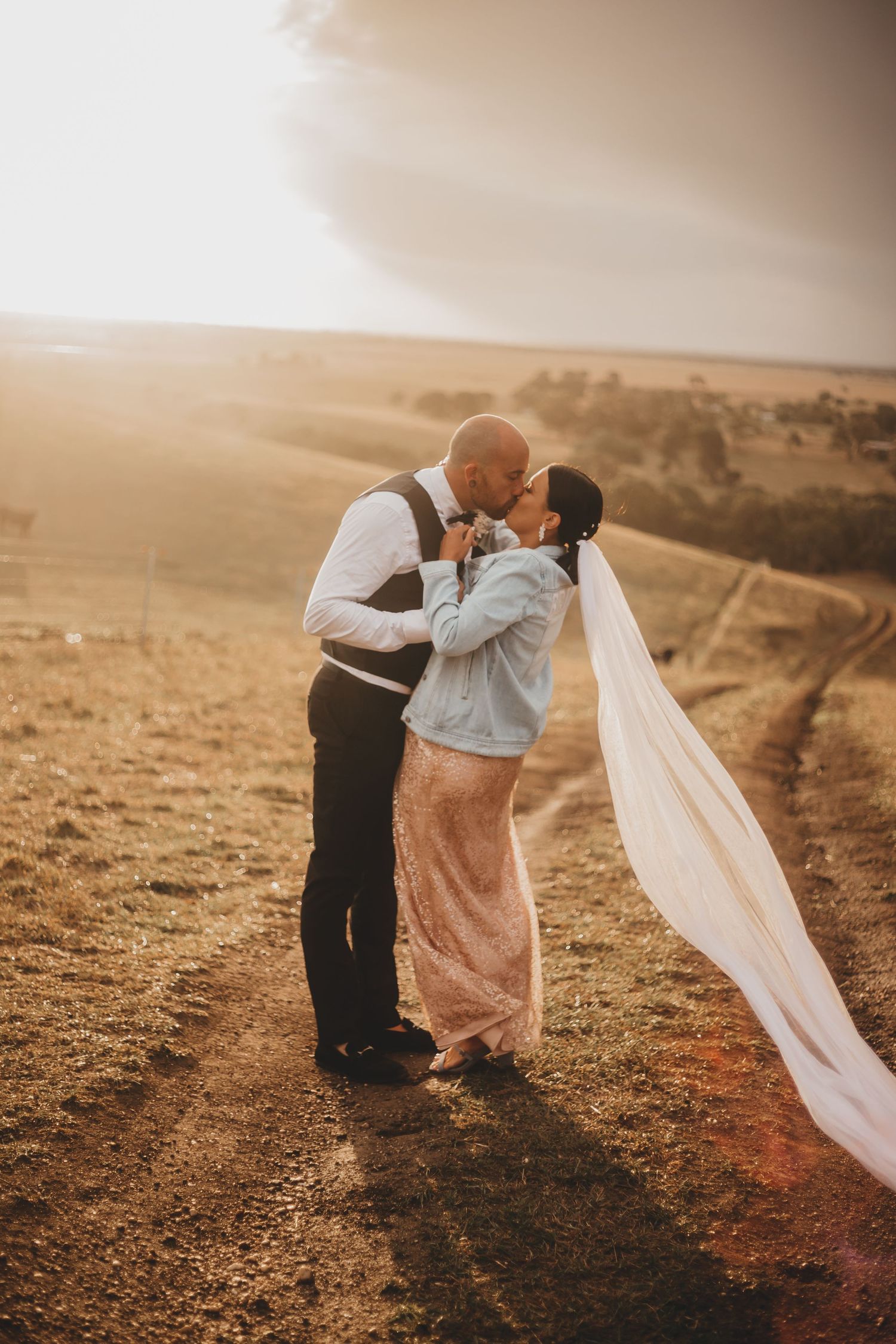 A couple shares a romantic moment at sunset on a hilltop, with the bride wearing a flowing veil and denim jacket.