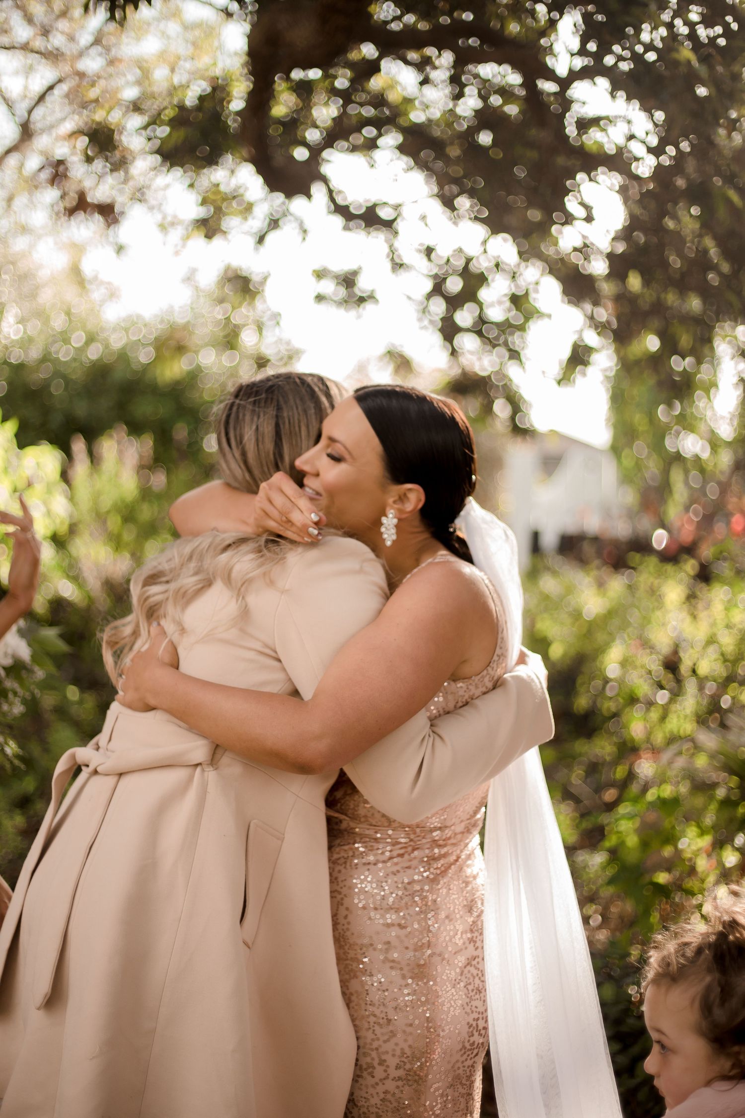 Two people share an emotional embrace under dappled sunlight in a garden setting with a white veil and rose gold dress.