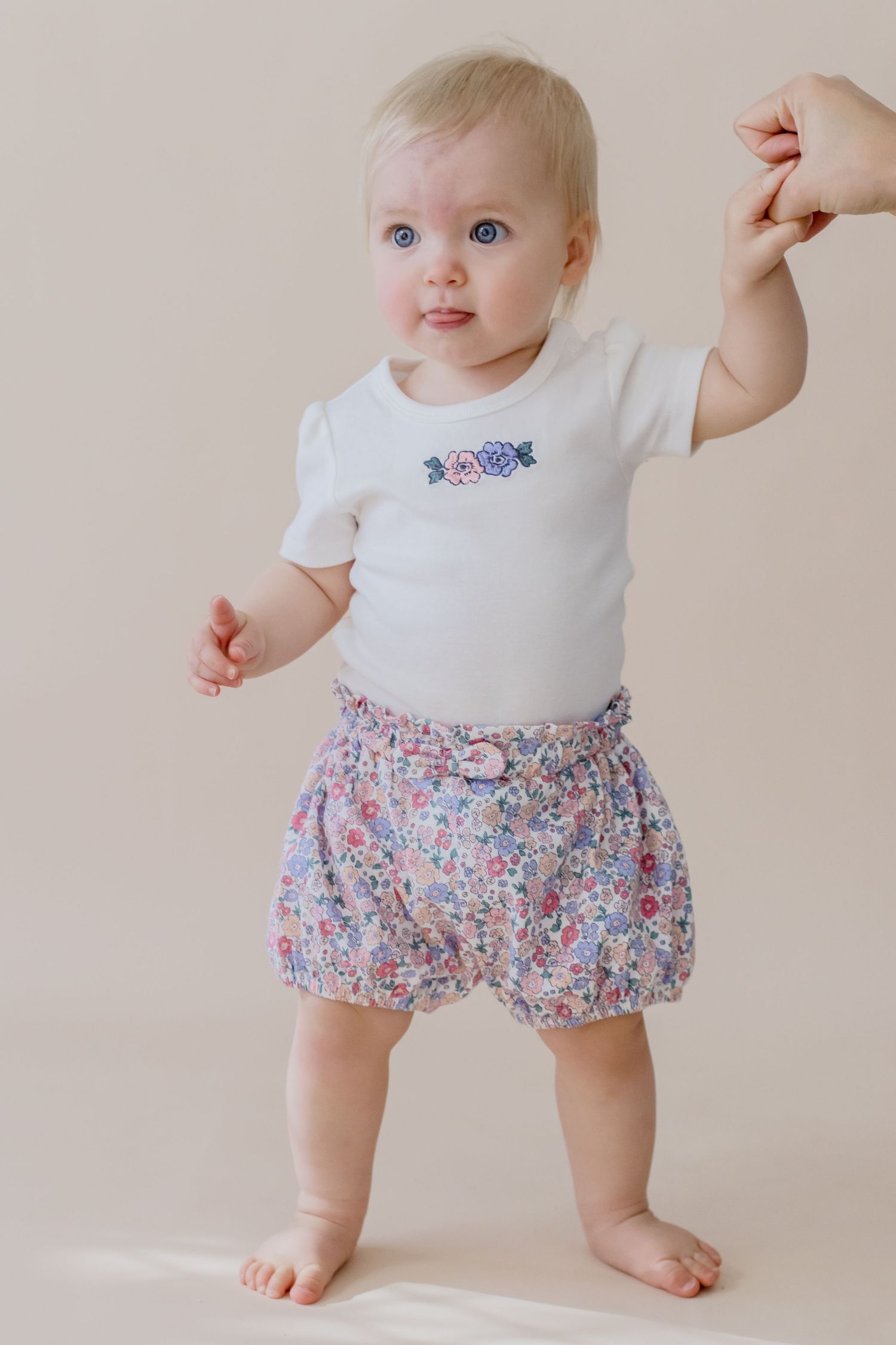 Baby wearing white onesie and floral bloomers stands barefoot against neutral background holding someone's hand.