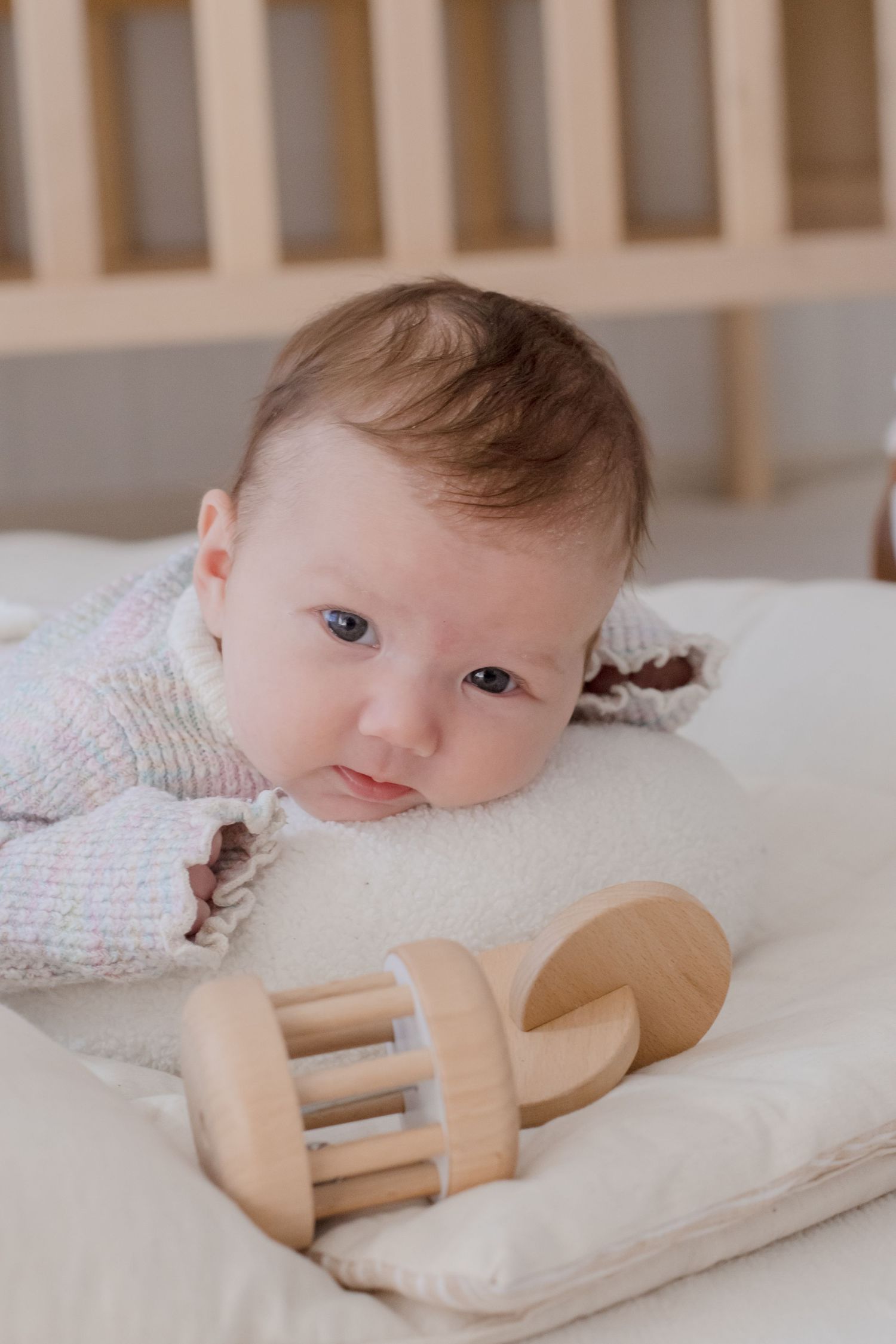 Baby laying on white bedding reaching for a wooden teething toy.