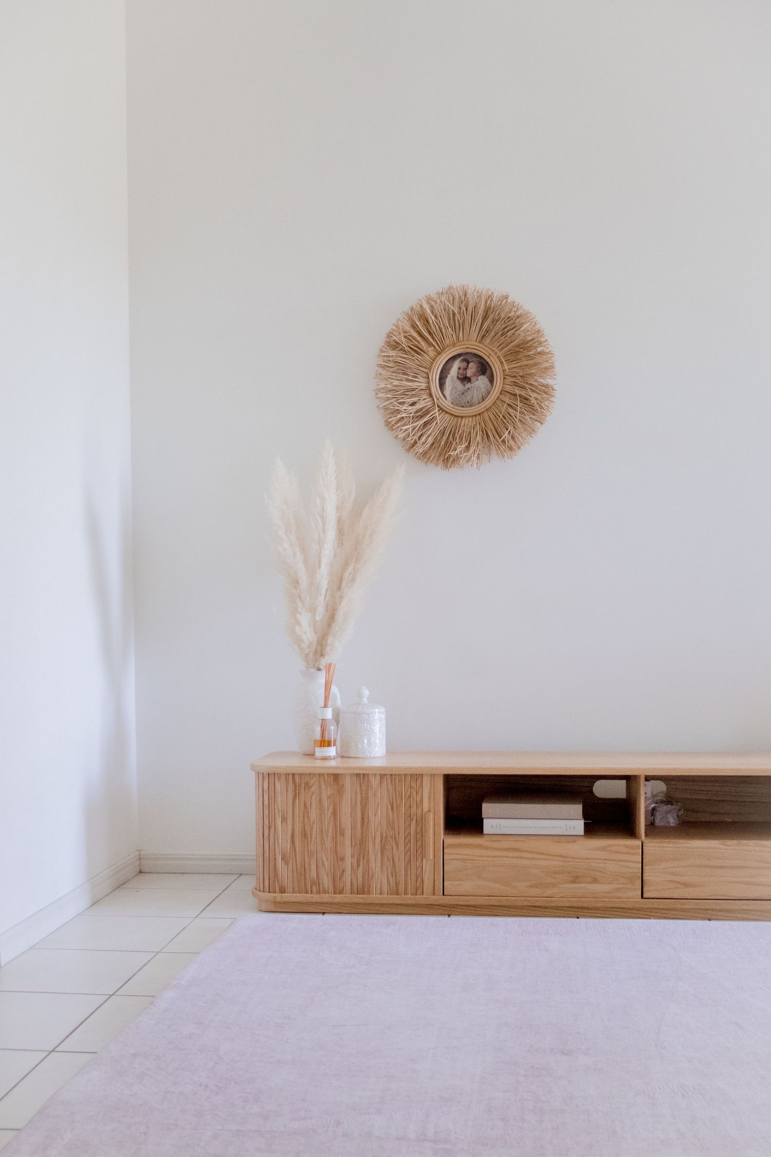 Minimalist living room with wooden TV unit, circular jute wall decor, and pampas grass in a glass vase.