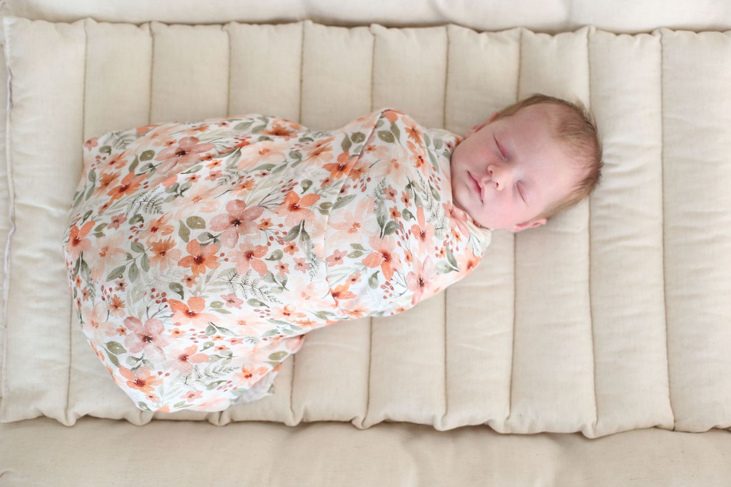 Sleeping newborn wrapped in floral swaddle blanket on cream colored cushion.