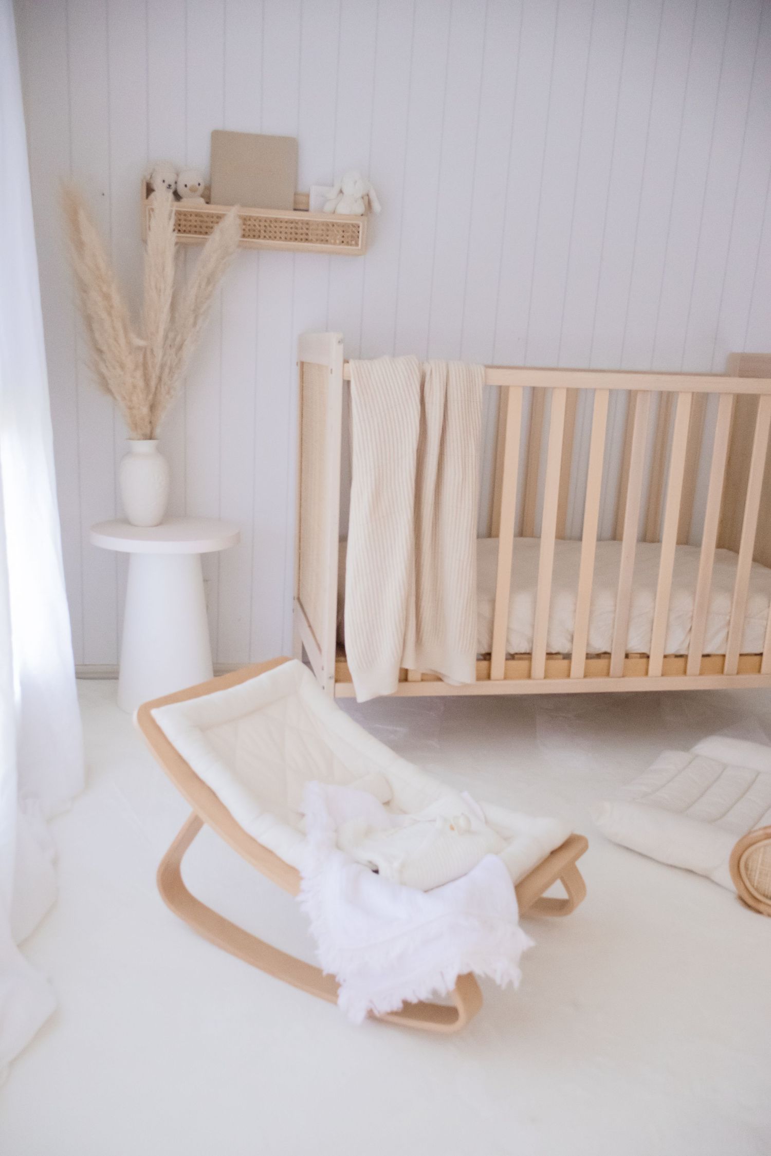 Minimalist nursery with wooden crib and rocking chair in white room with pampas grass decor.