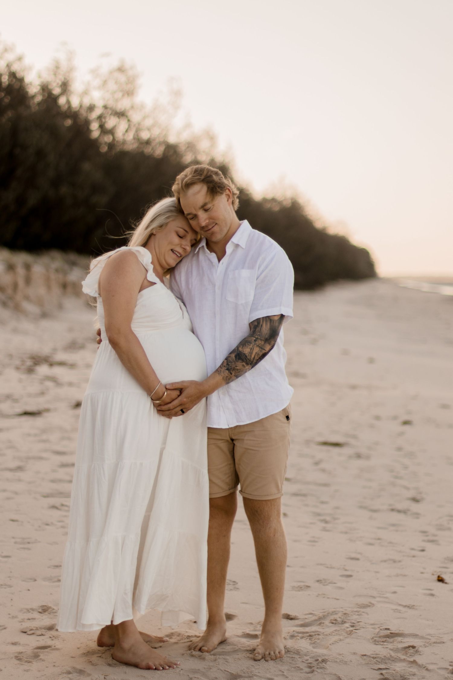 A romantic maternity photoshoot shows a couple embracing on a sandy beach at sunset.
