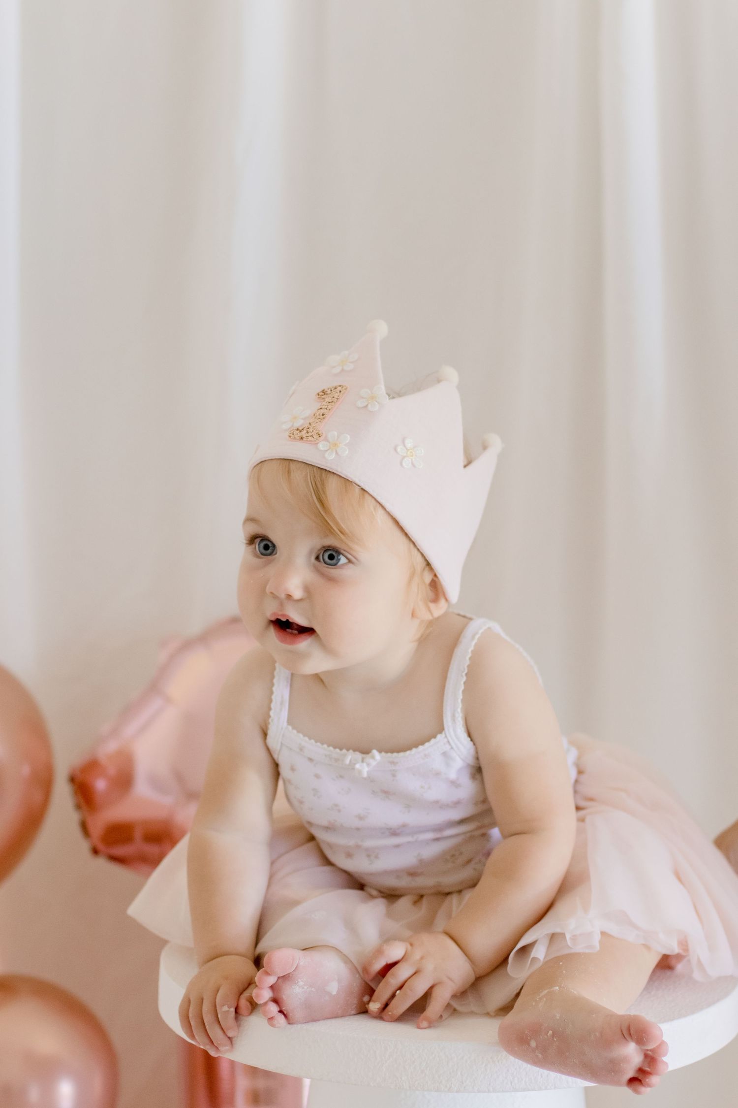 A cheerful baby in pink tulle dress and crown poses on white pedestal with balloons.