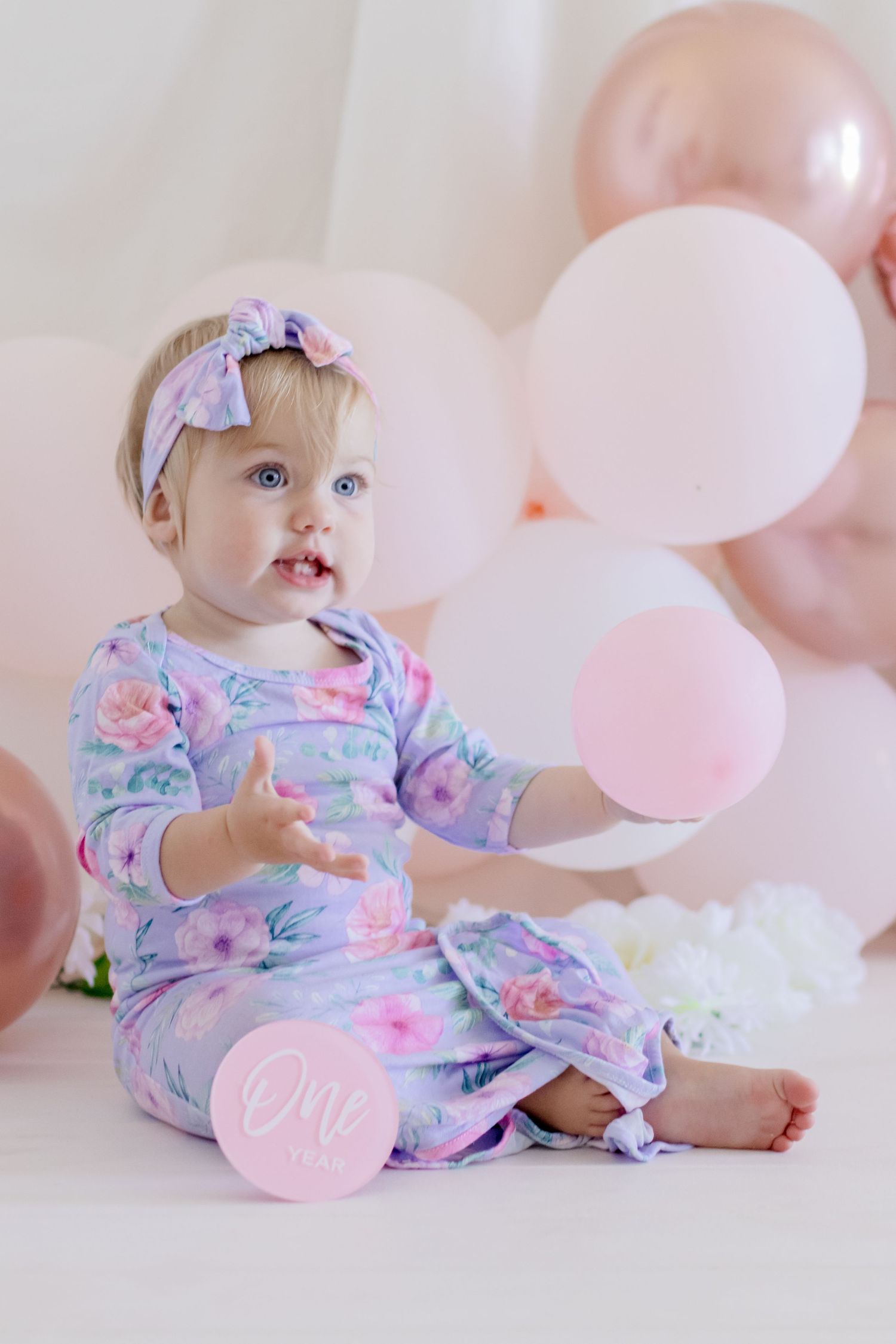 An infant in pastel floral sleepwear sits among pink and white balloons.