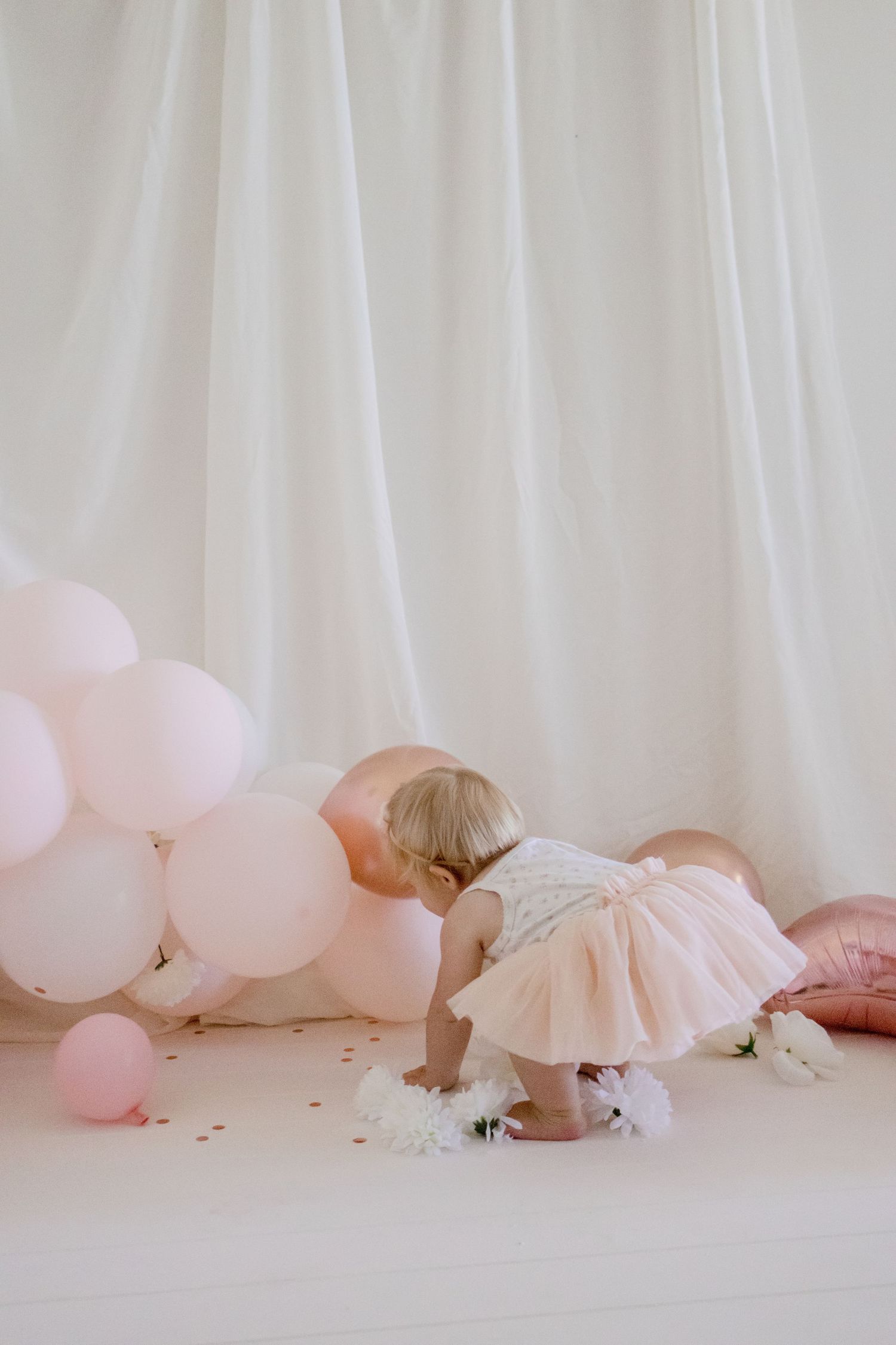 A toddler in a tulle dress bends down to explore decorative elements on a white floor.