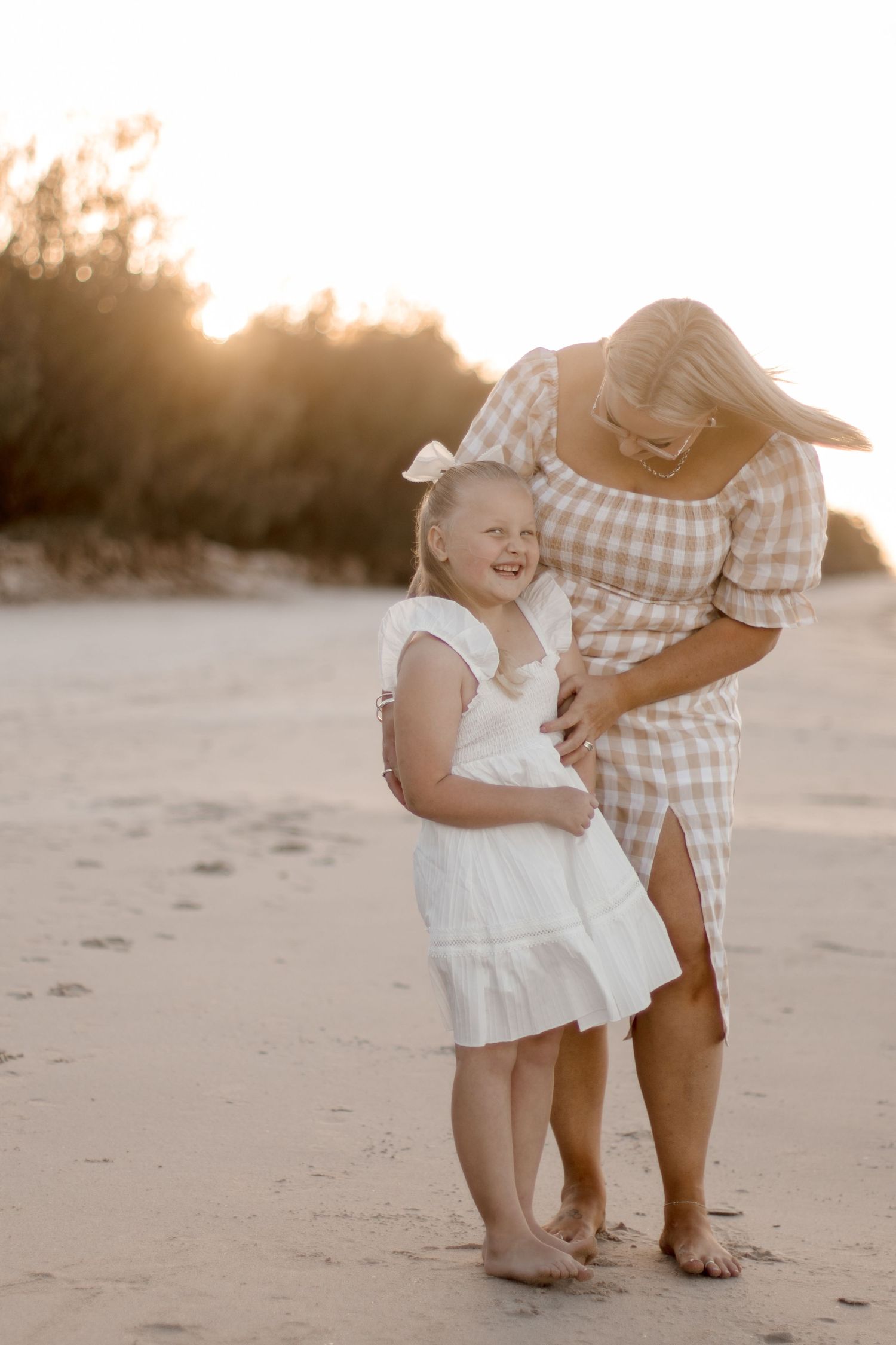 Two people share a joyful moment while standing barefoot on a sandy beach at sunset in matching light colored outfits.