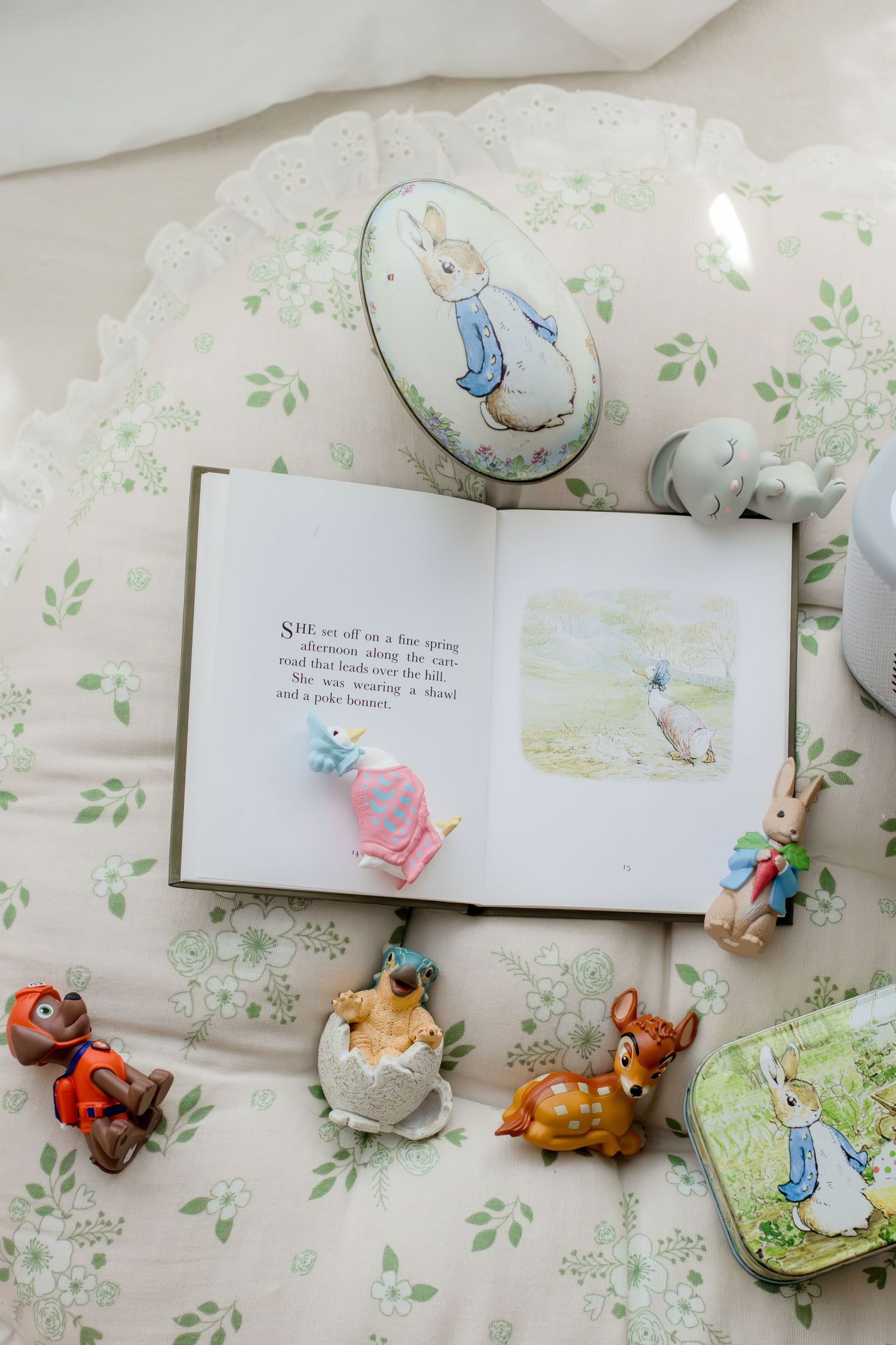 Open Peter Rabbit book surrounded by vintage figurines, Easter eggs, and collectibles on floral bedding.