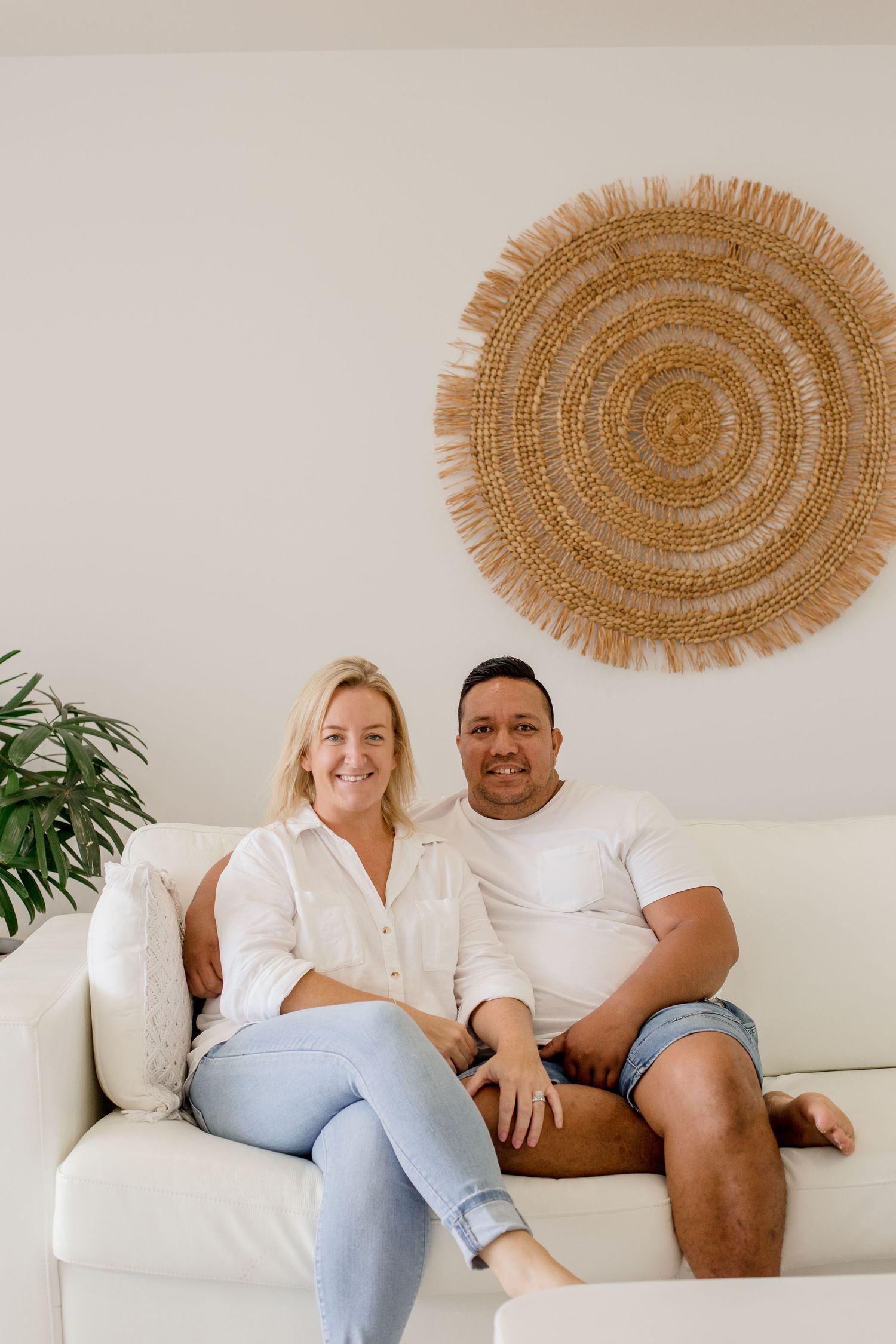 Couple sits close together on white couch under circular woven wall decoration.