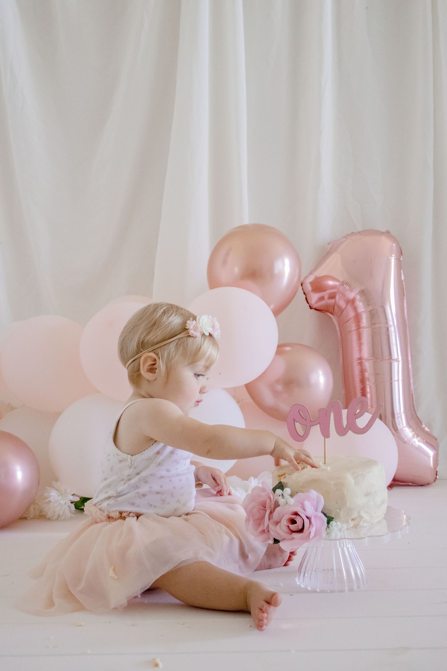 Baby in white dress exploring birthday cake setup with pink balloon decorations.