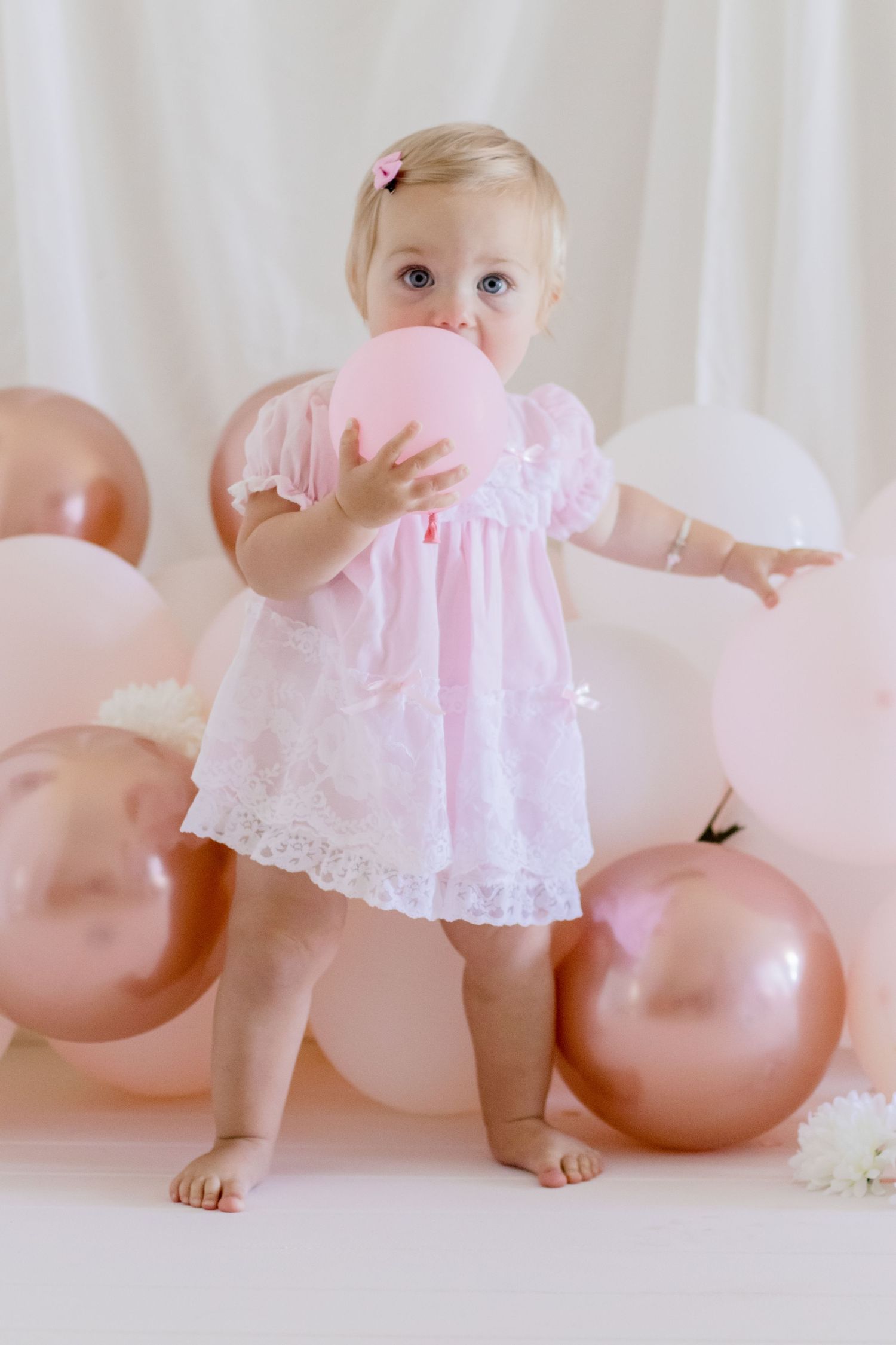Baby in lace dress holds pink balloon against white backdrop with rose gold decorations.