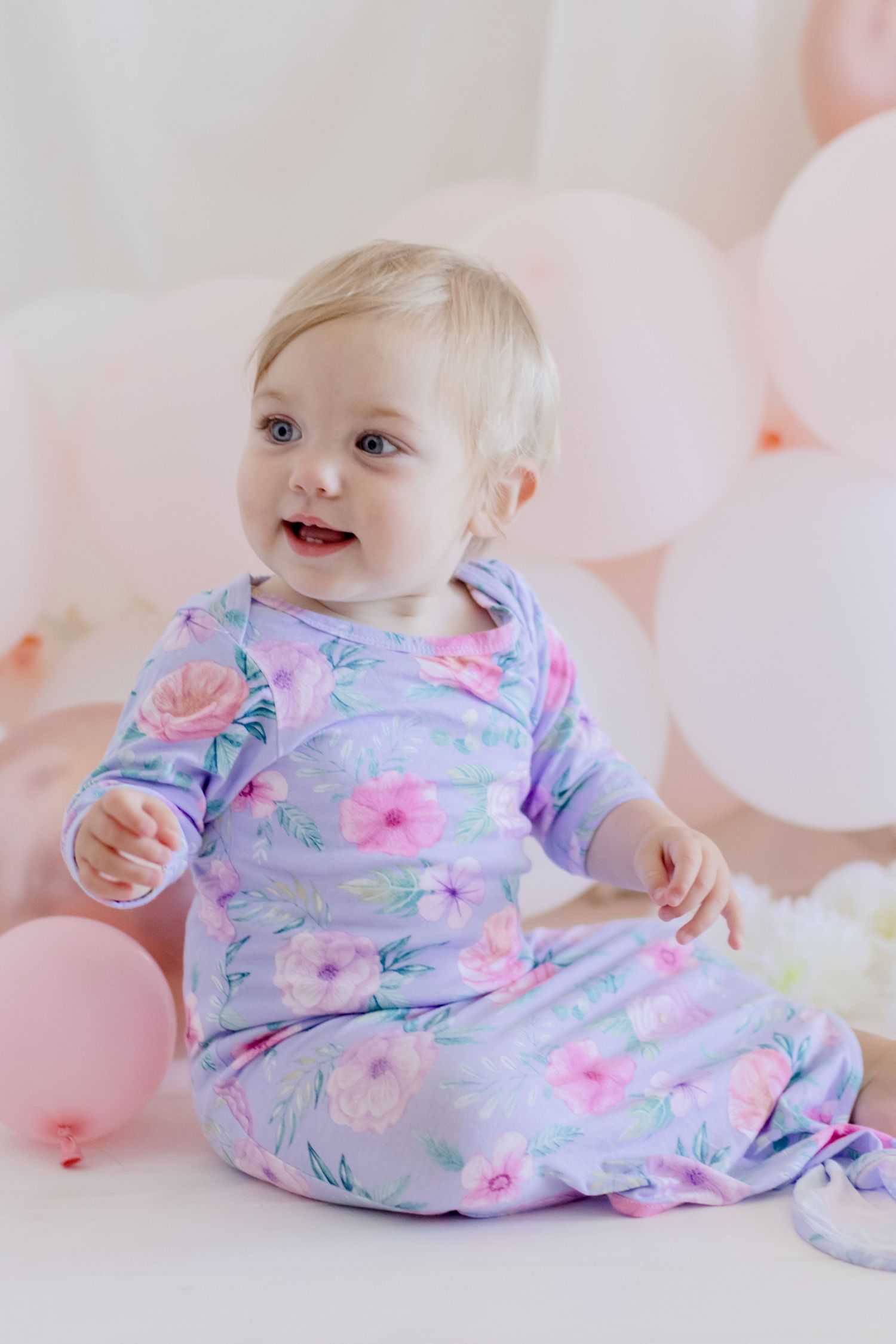 Smiling baby in pastel floral sleepwear against pink balloon backdrop.
