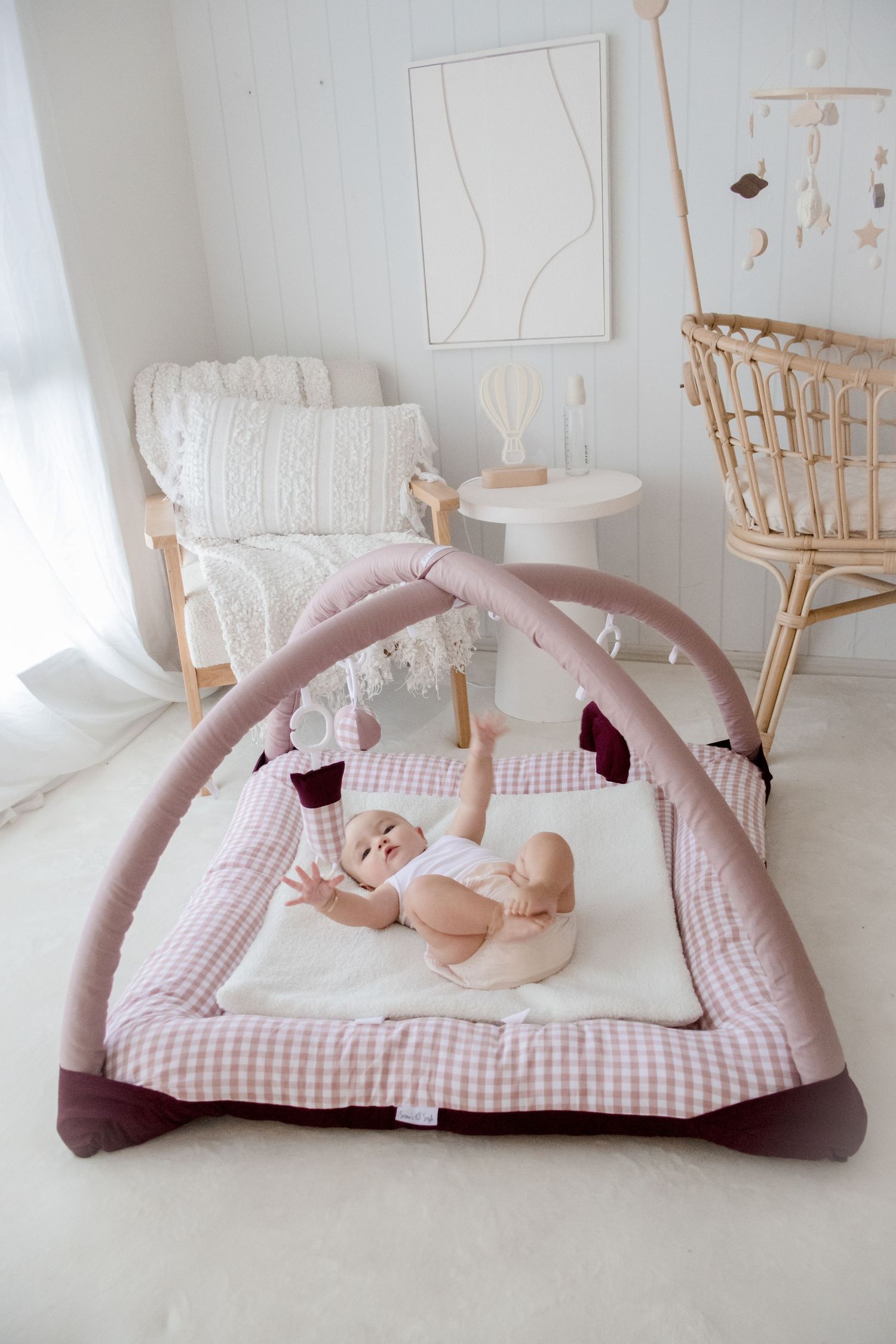 Baby activity gym with pink arches and striped cushioned mat in a minimalist nursery with white decor and rattan furniture.