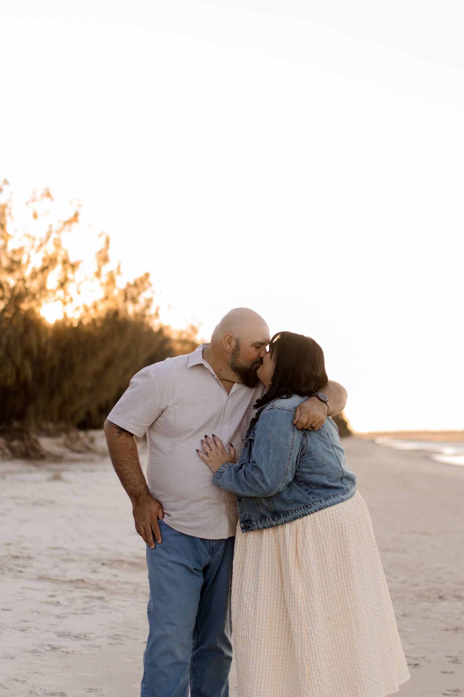 A romantic sunset beach portrait of a couple embracing in casual attire on sandy shores at golden hour.
