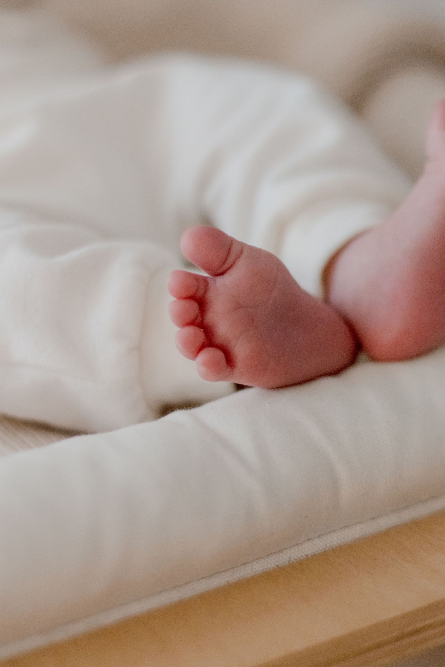 Tiny newborn baby feet peek out from soft white bedding on a wooden surface.