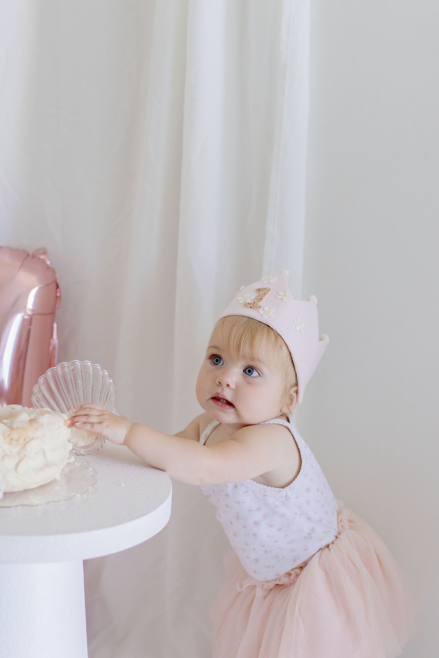 A toddler in pink tulle dress and crown reaching up to white party table.